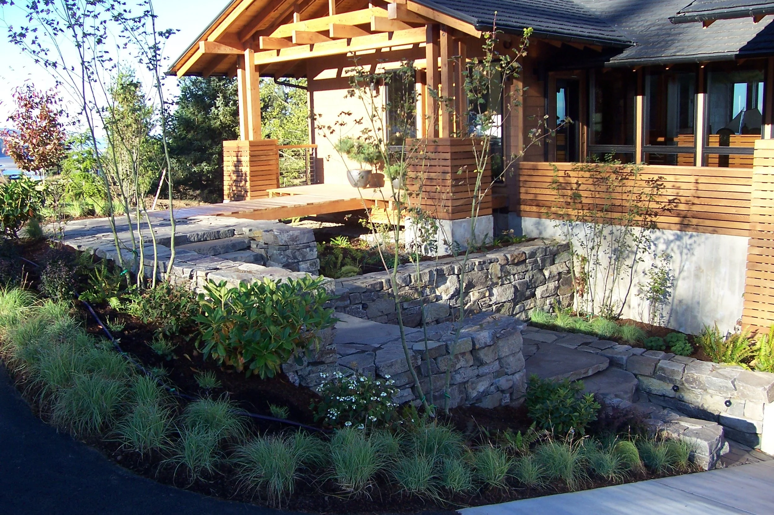 A house with a wooden deck and a sloped roof on the Oregon Coast, surrounded by a landscaped garden with stone pathways, various green plants, shrubs, and trees.