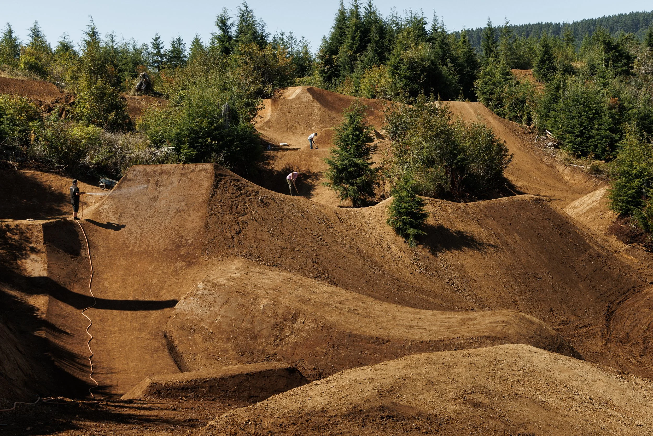 People working on a dirt bike trail in a forested area with dirt jumps and ramps.