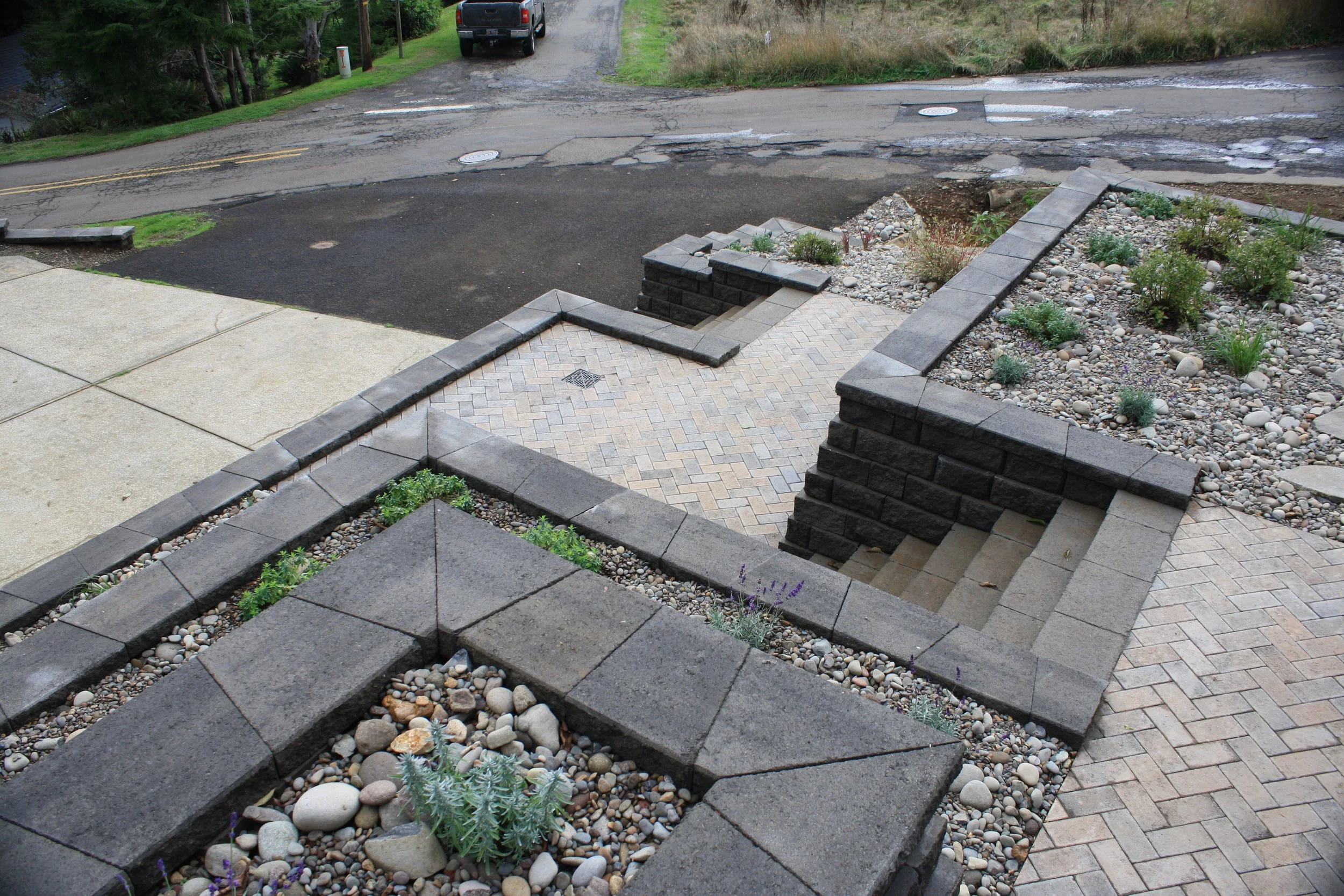 A front yard on the Oregon Coast with a brick walkway and stairs leading down to a sidewalk, with rock and soil landscaping on either side. The street beyond is cracked and is partially paved, with a black pickup truck parked on the side.
