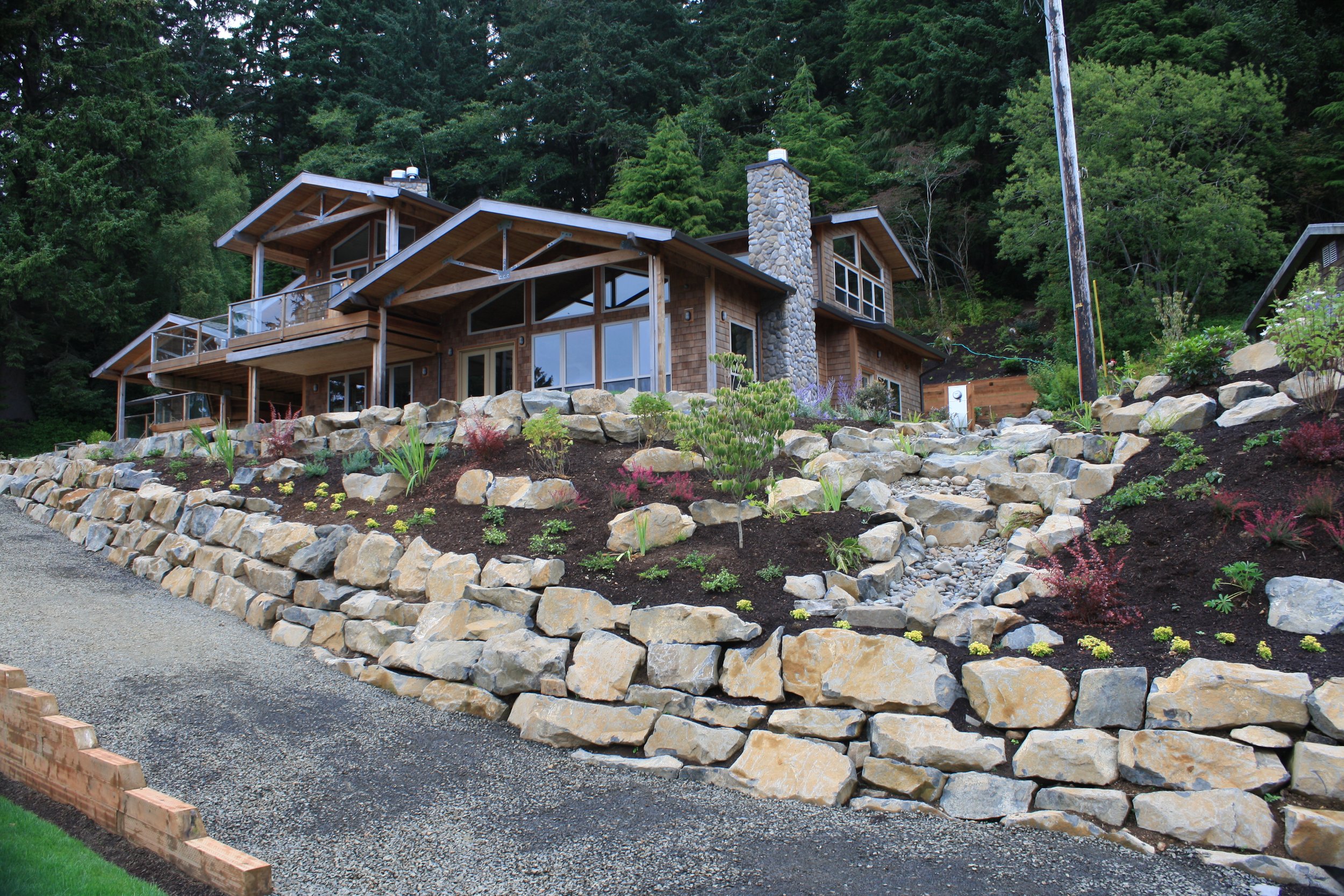 A modern house built on a slope on the Oregon Coast, a stone chimney, and wooden siding, surrounded by a landscaped yard with rocks, herbs, and flowers, and dense trees in the background.