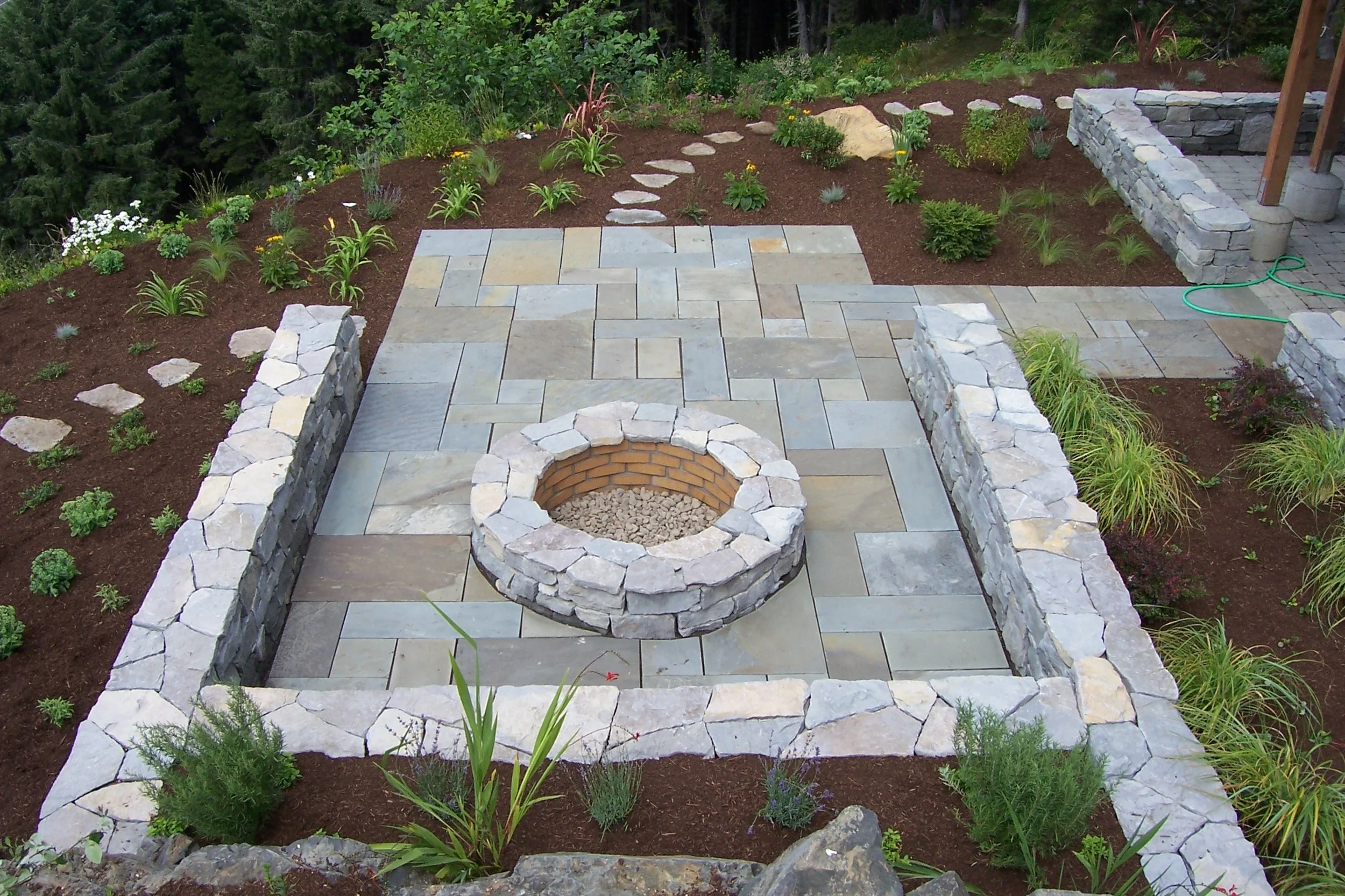 A landscaped backyard patio on the Oregon Coast with stone pavers and a circular fire pit, surrounded by flower beds with various plants and a green garden hose on the ground.