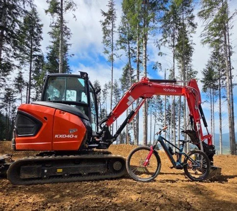 A small orange excavator with a red arm is lifting a mountain bike with a black frame, blue accents, and thick tires on a dirt area with tall trees in the background.