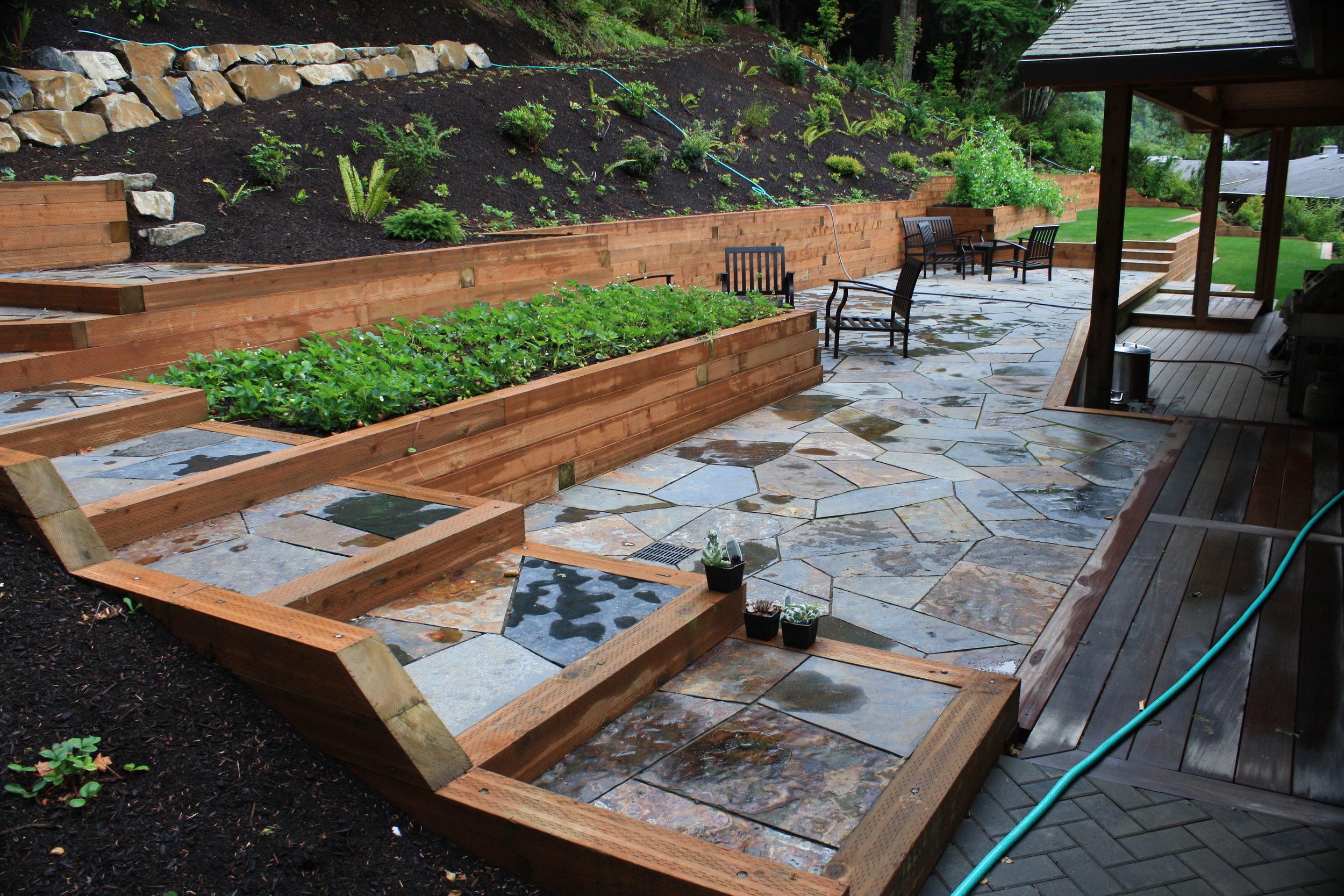 A backyard patio on the Oregon Coast with stone flooring and wooden planters filled with plants and young greenery, backed by a sloped garden hillside with shrubs and a stone retaining wall.