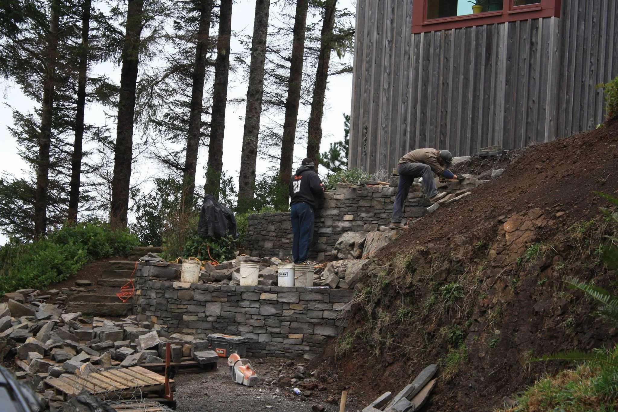 Two construction workers building a stone retaining wall on a hillside near a house with a red exterior and large windows. The workers are using tools, with buckets and construction materials around them, and tall trees in the background.