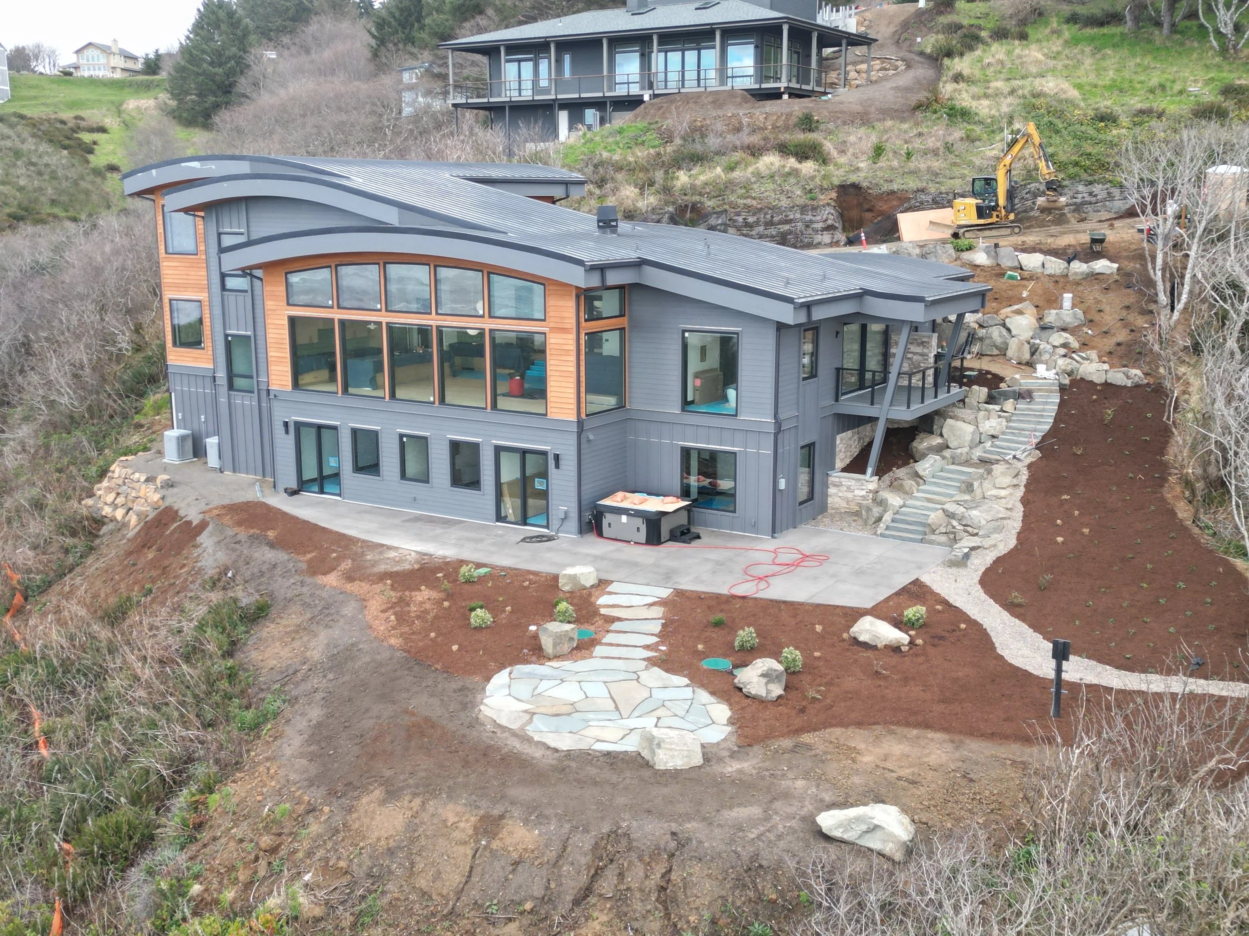New house under construction on a hillside on the Oregon Coast, with large windows and curved roof, construction equipment working behind it, and a partly landscaped yard with stepping stones and small plants.