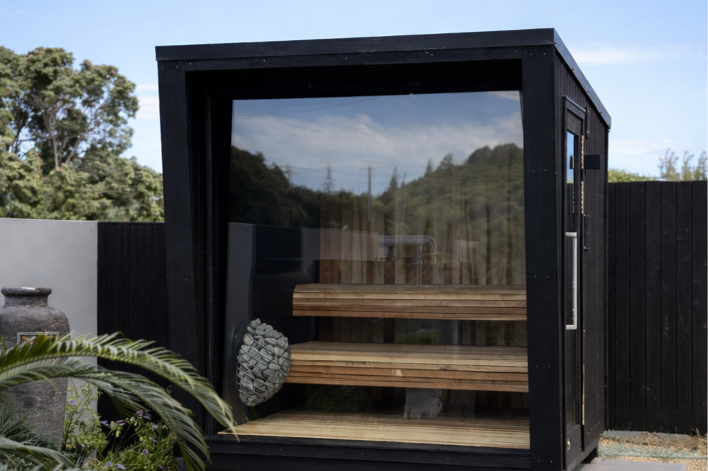 An outdoor wooden sauna with a glass front door and horizontal wooden slats, situated in a garden with green plants and trees nearby.