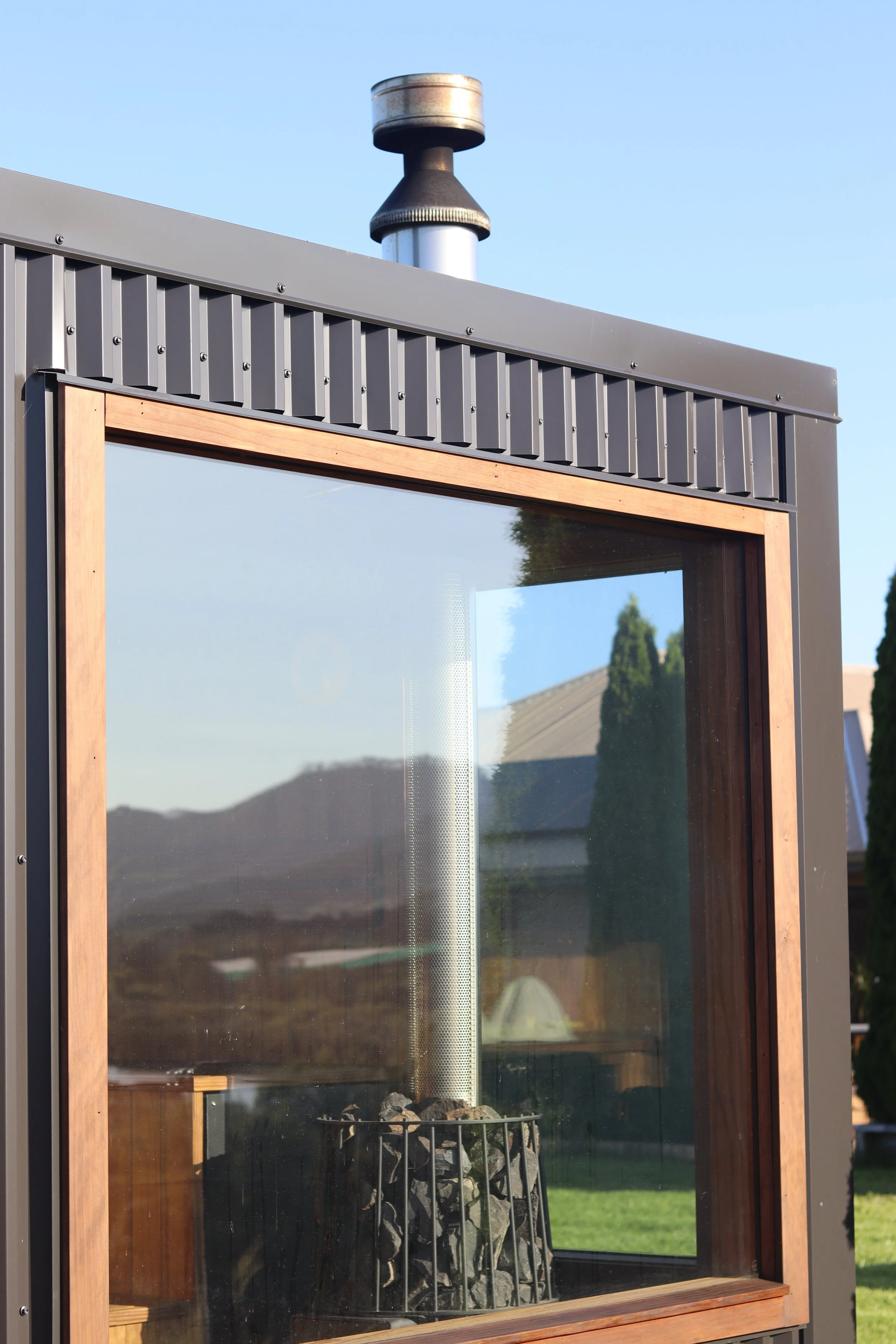 A modern outdoor fireplace with a glass enclosure and wood trim, showing a chimney pipe, metal siding, and a grassy backyard with trees and houses in the background.