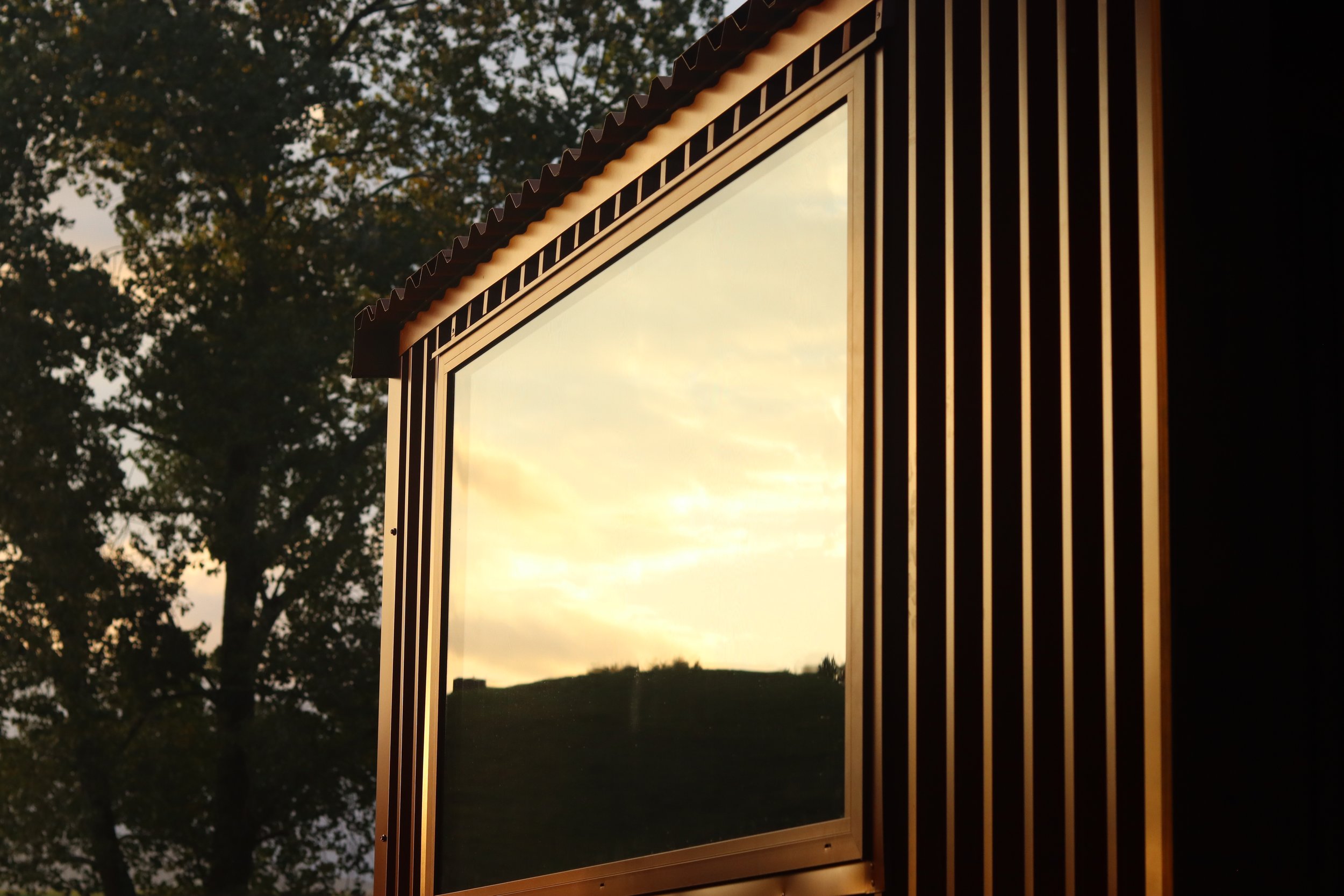 A large rectangular mirror with a decorative frame featuring vertical lines, reflecting a cloudy sky with a hint of sunset, set outdoors with a tree in the background. Home Sauna - Custom-built, handcrafted sauna made in New Zealand