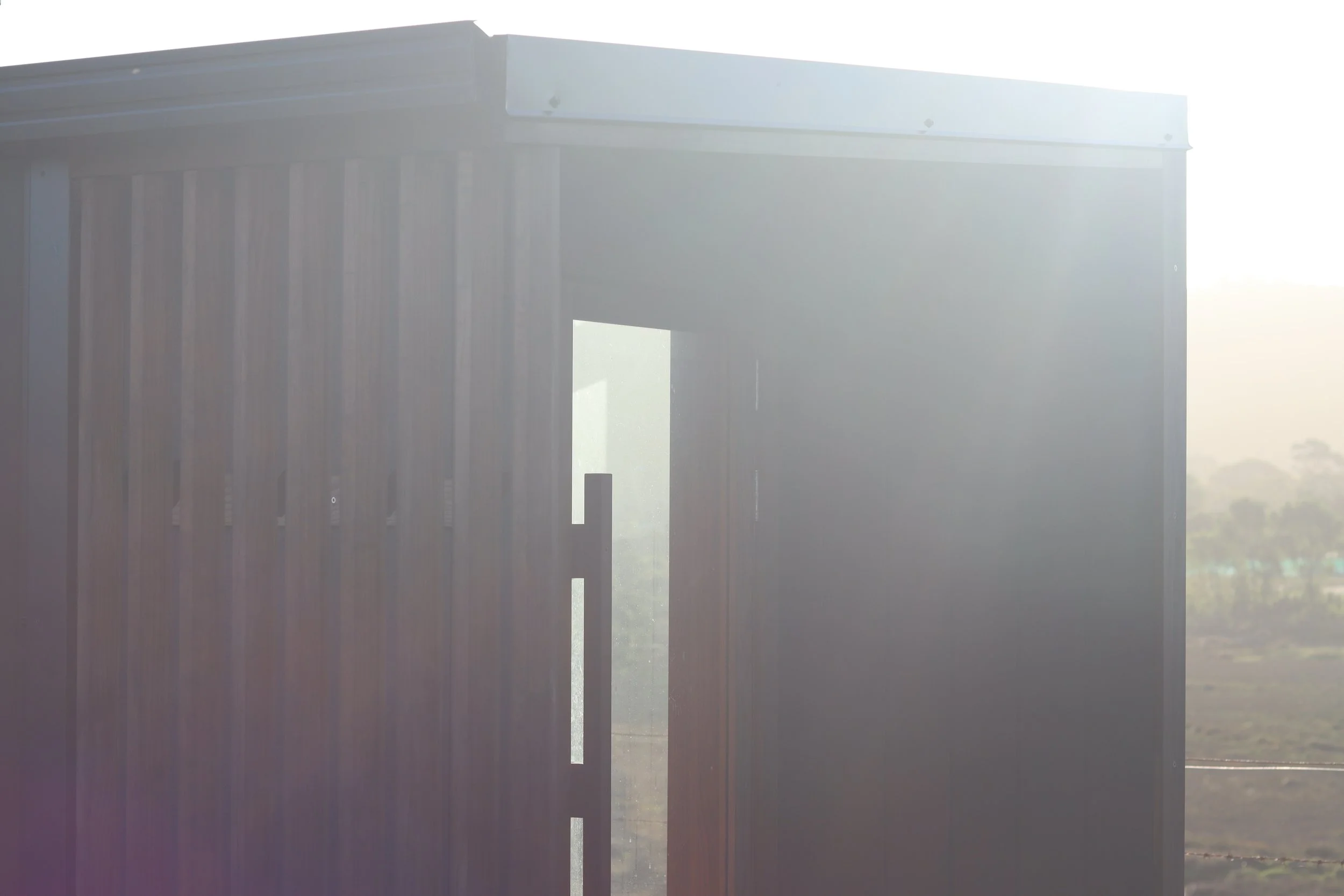 A modern wooden structure with vertical paneling and an open doorway, backlit by bright sunlight.