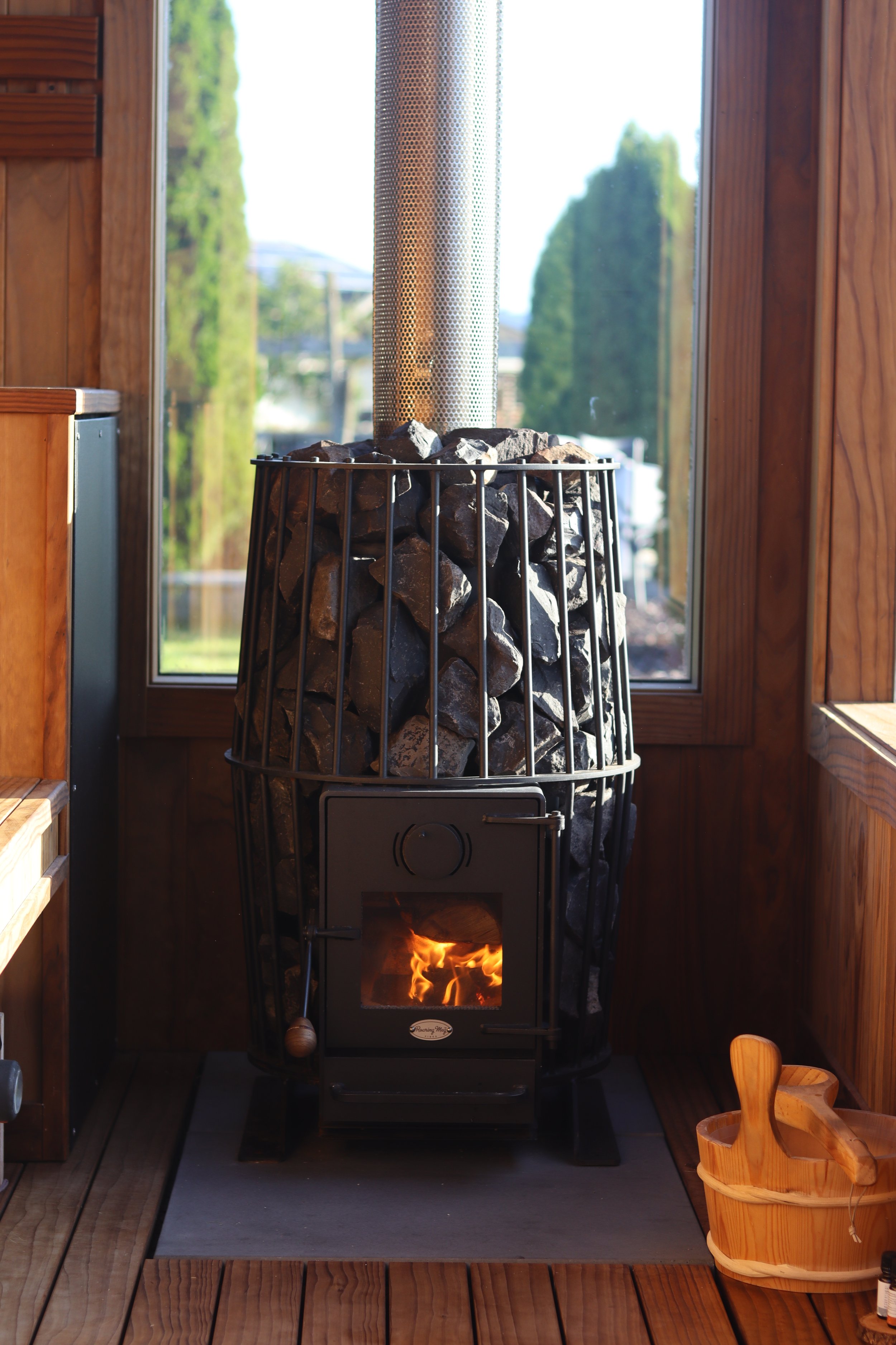 A sauna heater with rocks on top, inside a wooden room with a large window, burning fire visible through the door, and a wooden bucket with a wooden dipper beside it.