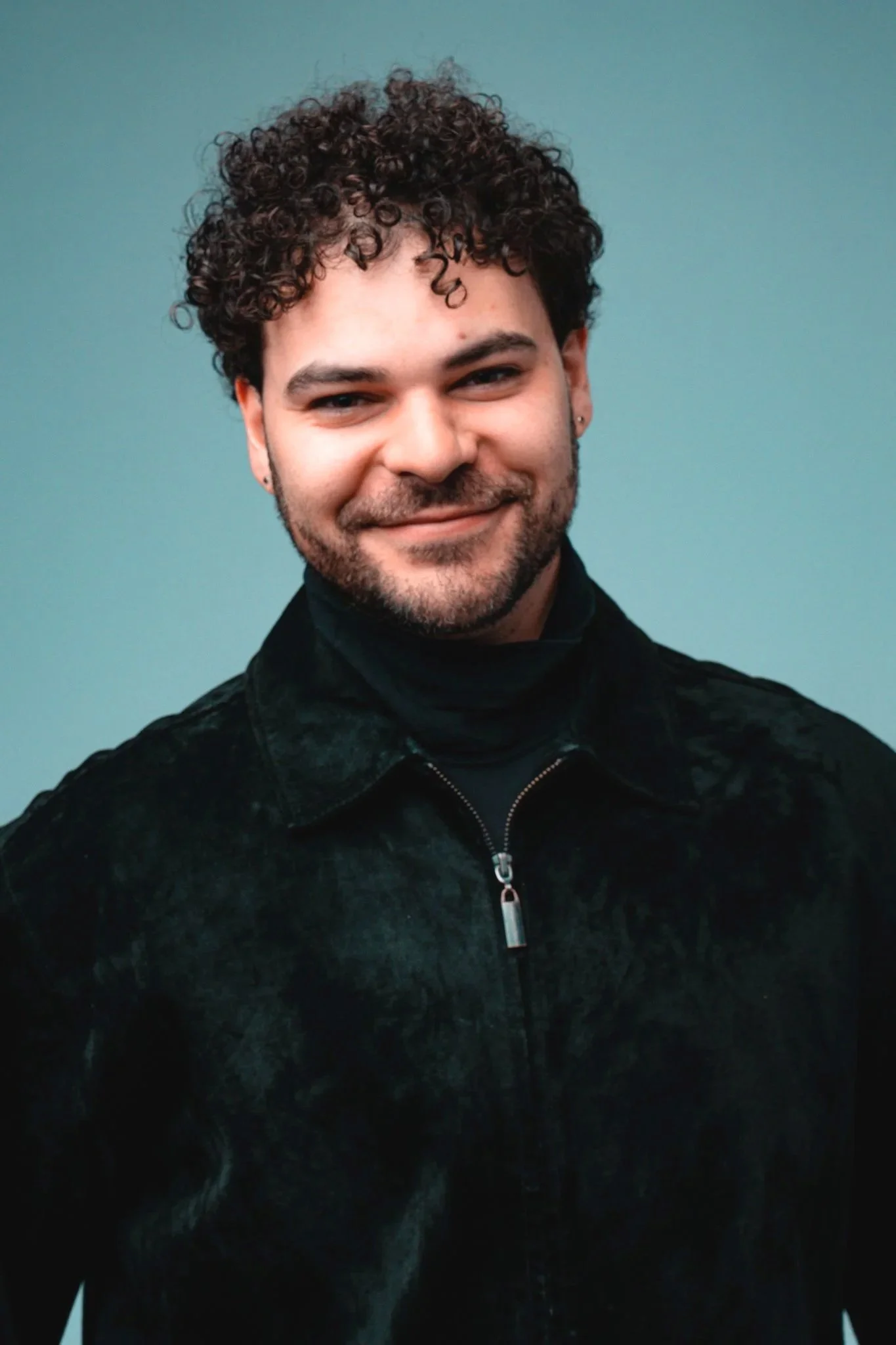 A young man with curly dark hair, beard, and earrings, smiling, wearing a black jacket and turtleneck against a plain background.