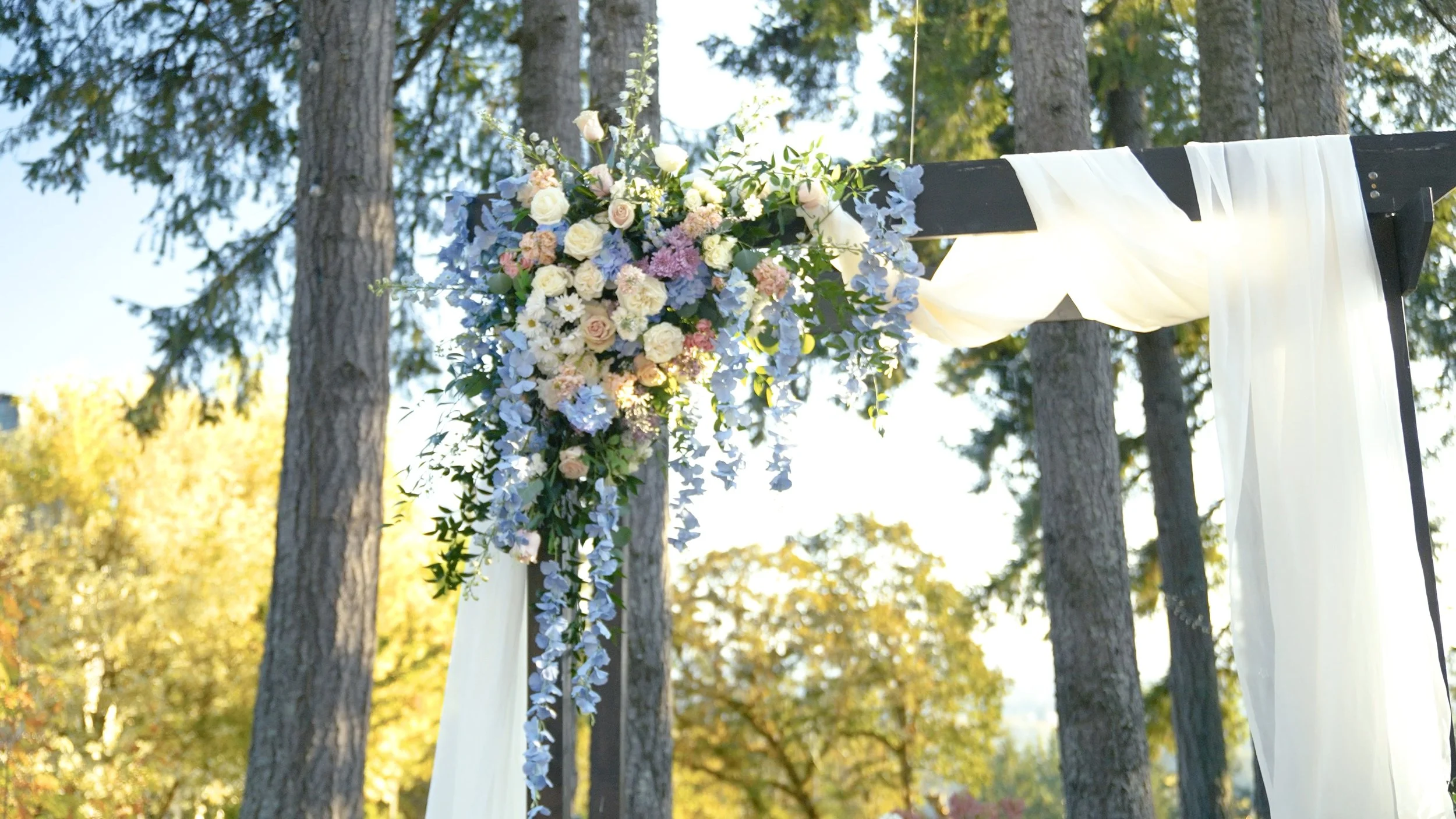 A wedding arch decorated with a bouquet of white, pink, and purple flowers, along with sheer white fabric, set outdoors among tall trees during daylight.