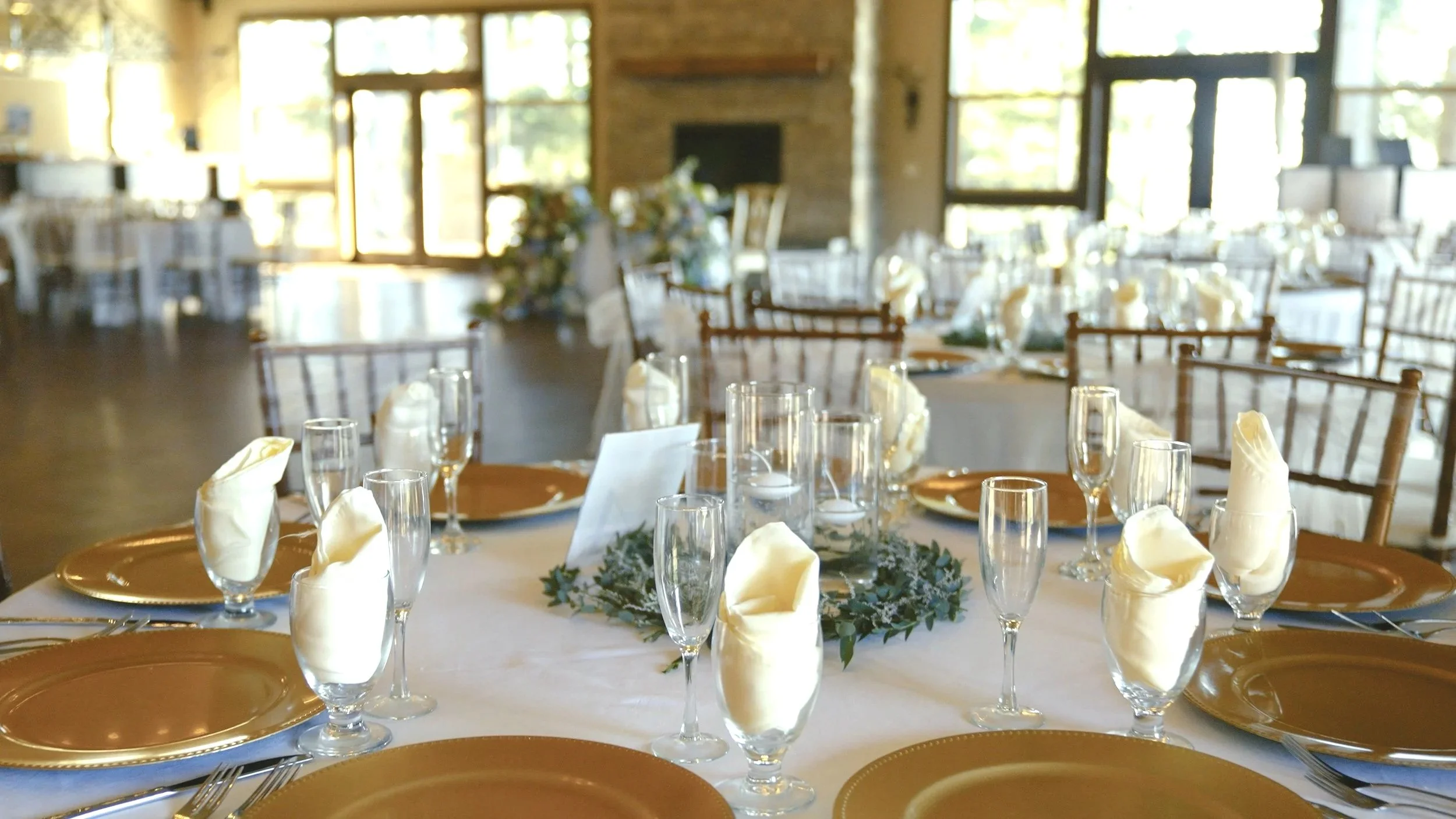 Elegant banquet table set with gold plates, champagne flutes with napkins, and floral centerpieces, in a bright room with large windows and wooden chairs.