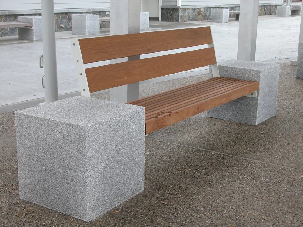 Empty park bench made of wood and metal, supported by two large concrete blocks, located on a concrete and gravel surface at a park or public space.