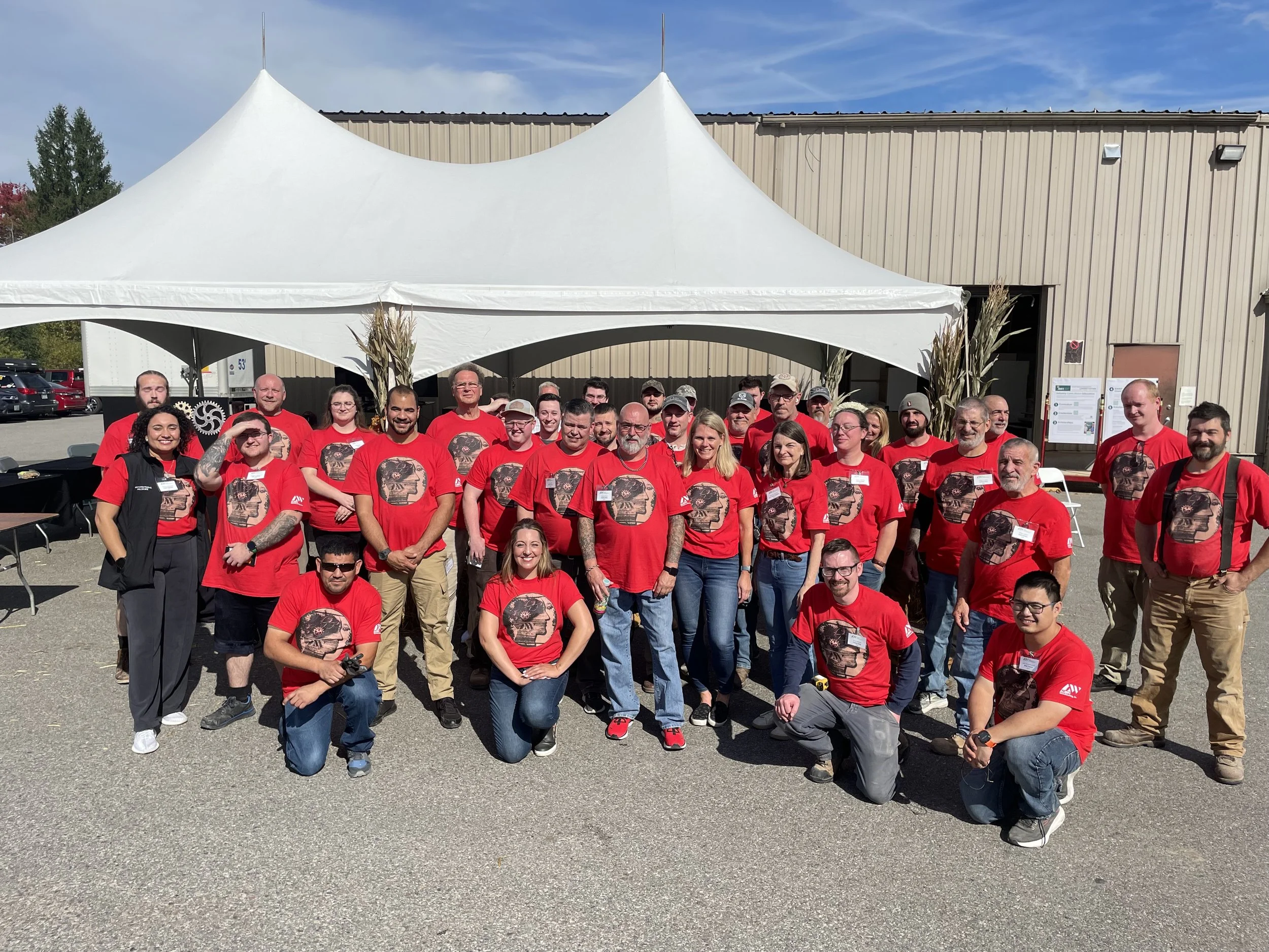 A large group of people, mostly wearing matching red T-shirts with a graphic design, are gathered outdoors in front of a building with beige vertical siding. They are posing for a group photo under a white canopy tent. The sky is clear and blue.