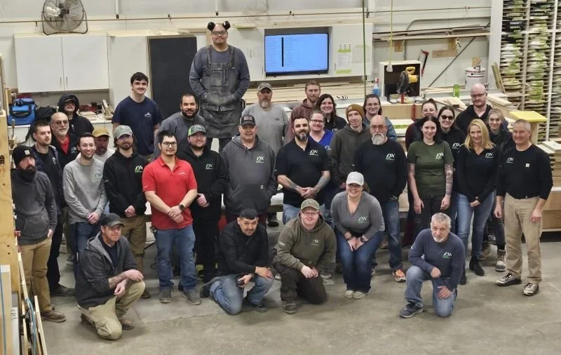 Group of 27 people posing together in a woodworking shop, some wearing logo shirts, with workbenches and tools in the background.