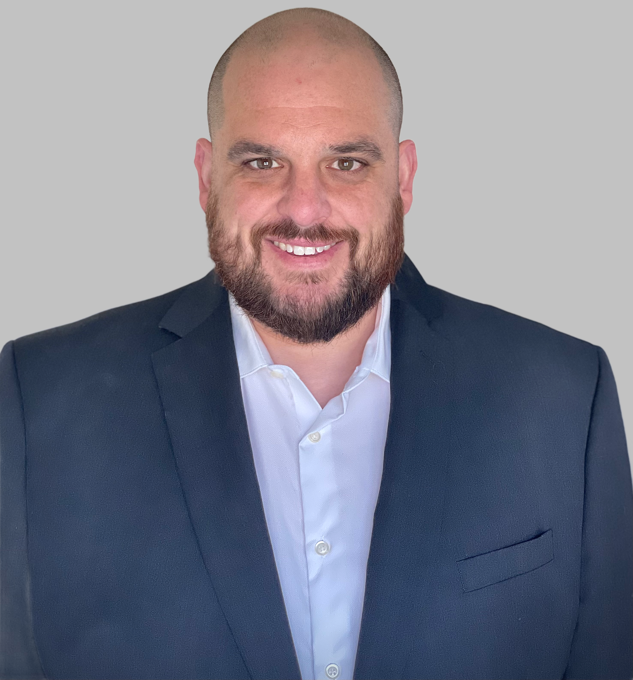 Headshot of a smiling man with a beard, wearing a white shirt and a dark blazer, against a gray background.