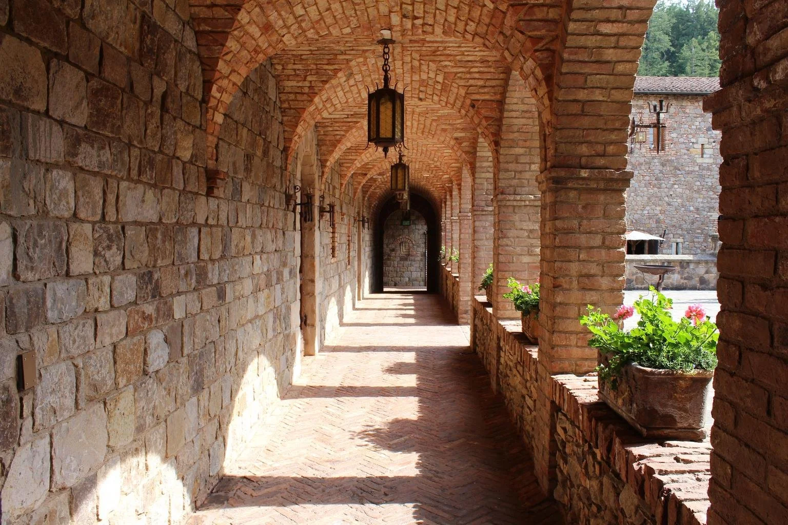 A covered brick corridor with hanging lanterns, potted plants, brick arches, and an open courtyard visible in the background.