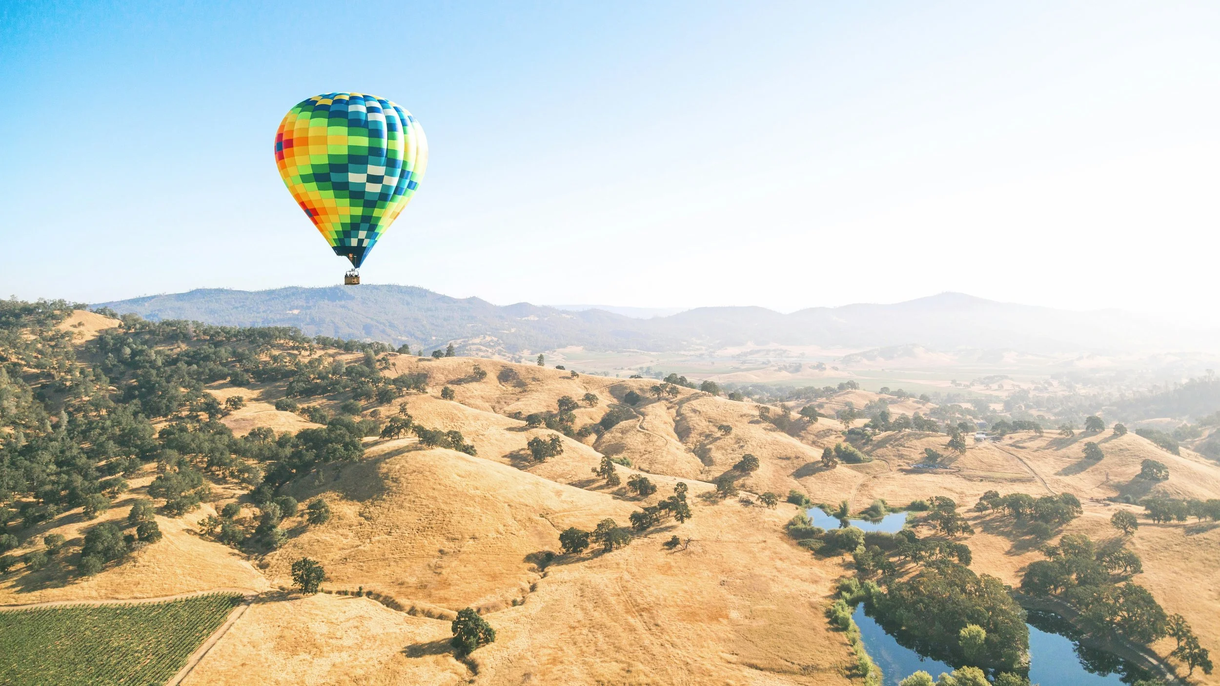 A colorful hot air balloon floating above a hilly landscape with trees and a small river, under a clear blue sky.