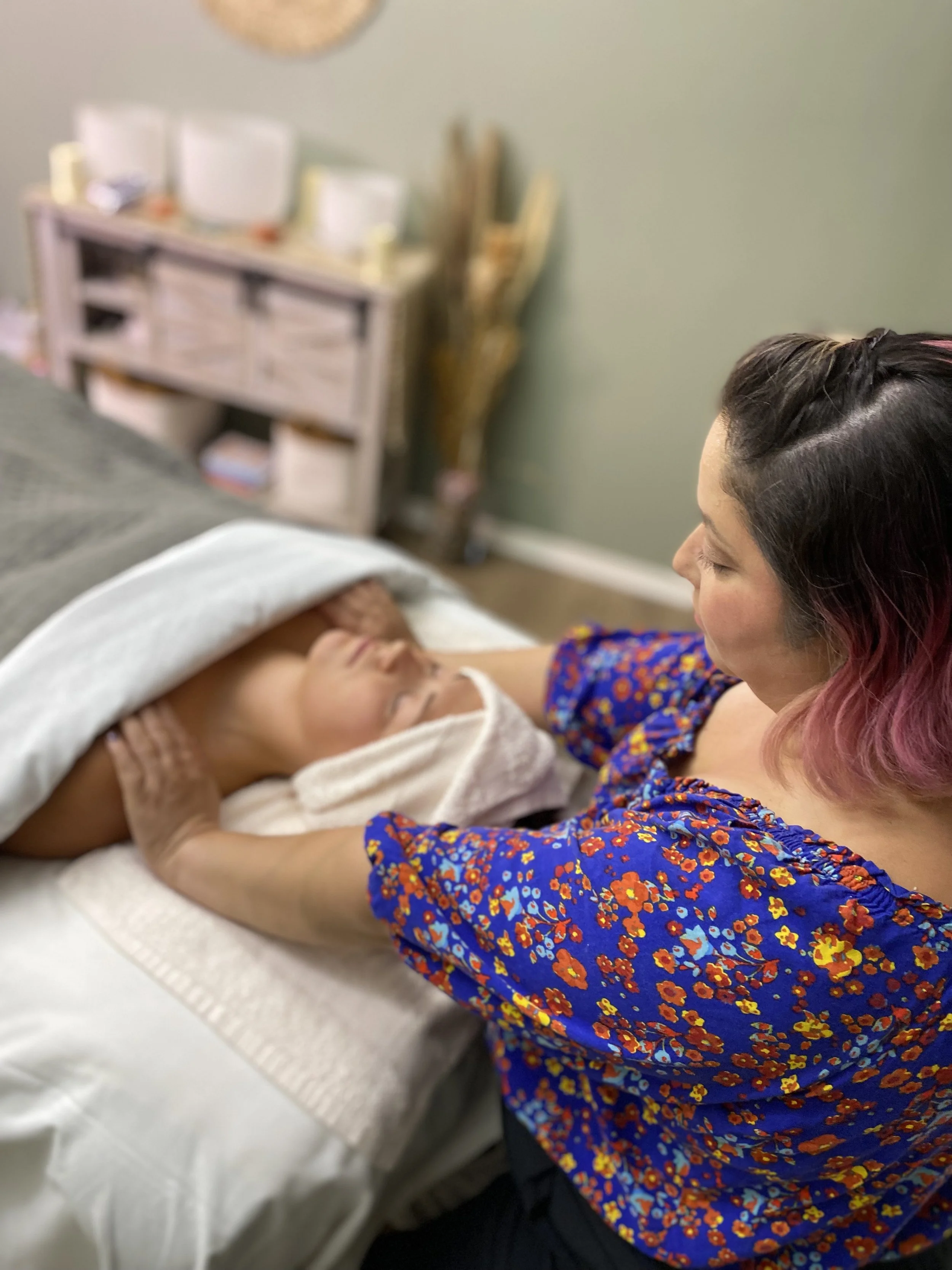 A woman with dark hair and pink tips, wearing a blue floral dress, is gently touching the forehead of a person lying on a bed, who appears to be resting with eyes closed and a towel on their forehead, in a room with a green wall and a blurry white cabinet in the background.
