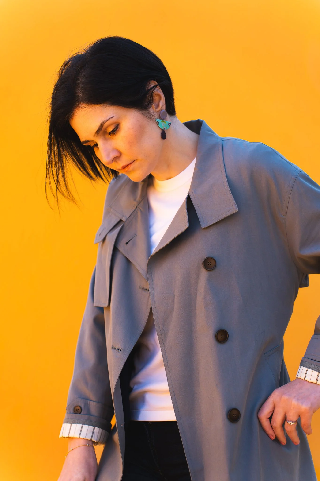 A woman with short black hair, wearing a grey trench coat over a white shirt, and colorful earrings, stands against a yellow background.