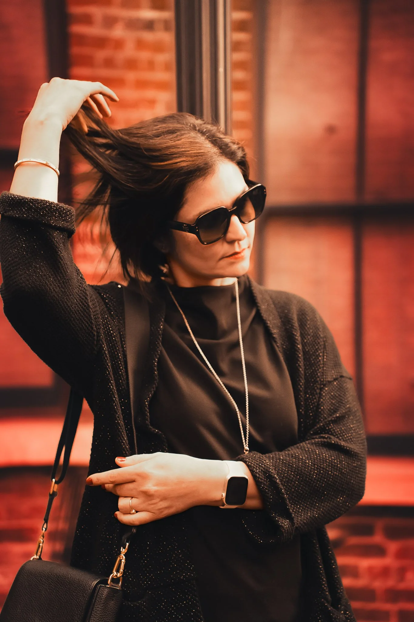 A woman with black hair wearing dark sunglasses, a black top, and a black cardigan, standing indoors against a brick wall background, holding her hair with one hand and crossing her arms, with a black purse, a silver necklace, and a smartwatch.