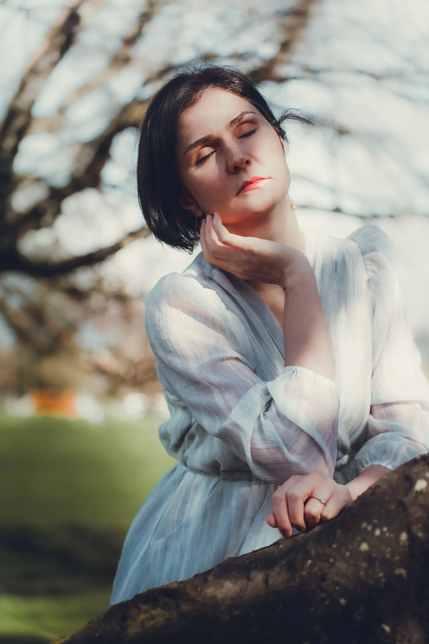 A woman with short dark hair, eyes closed, and a peaceful expression, resting her chin on her hand in an outdoor setting with blurred trees and green grass in the background.