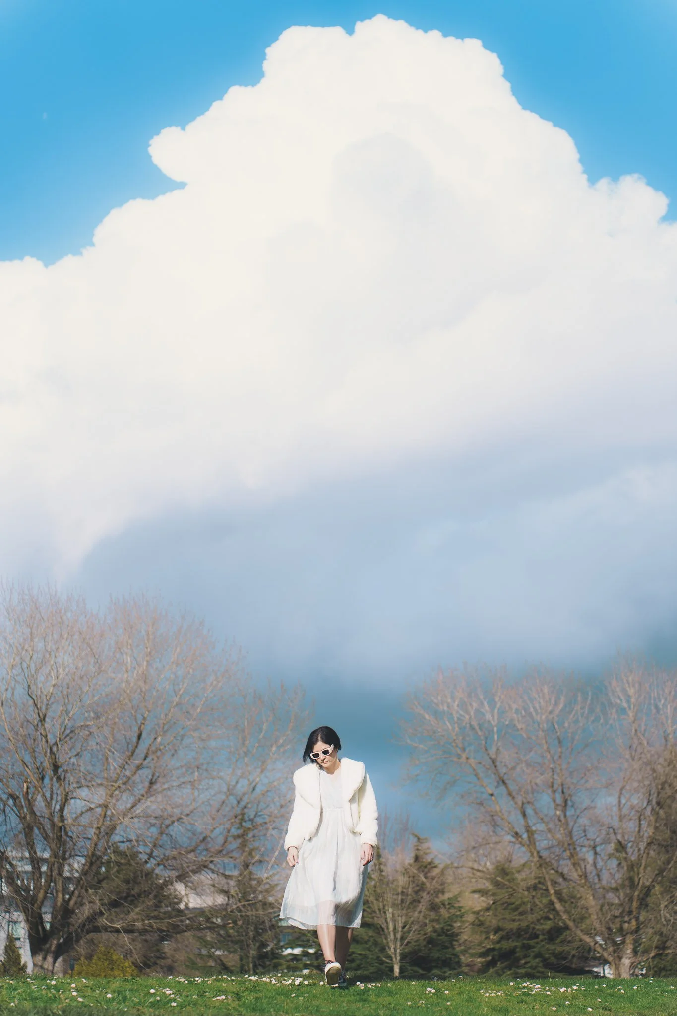 A woman in a white dress and coat walking on grass with bare trees and a large cloud in the sky.