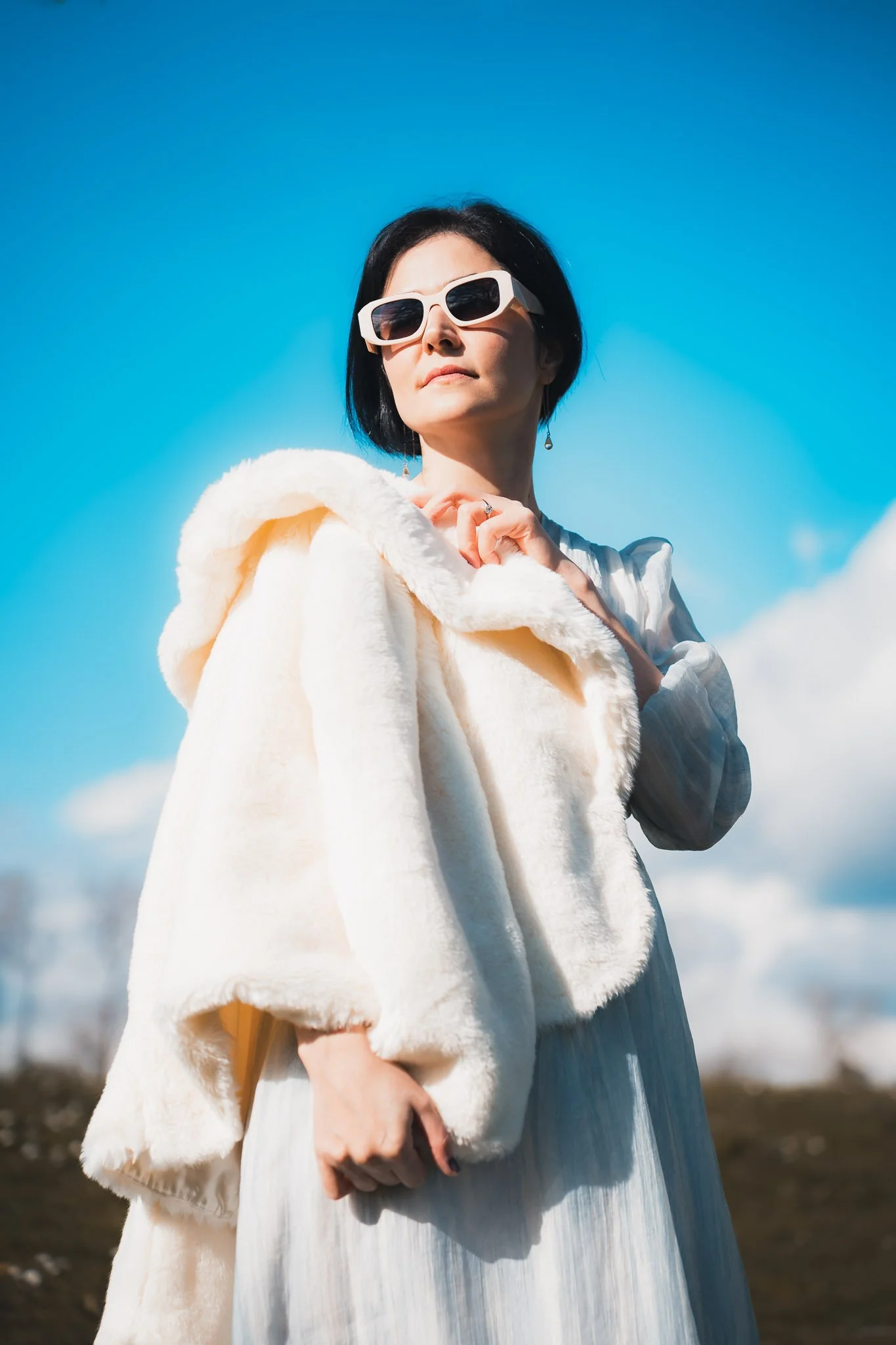 A woman with short dark hair and wearing white sunglasses holding a fluffy cream-colored coat outdoors against a blue sky with some clouds.
