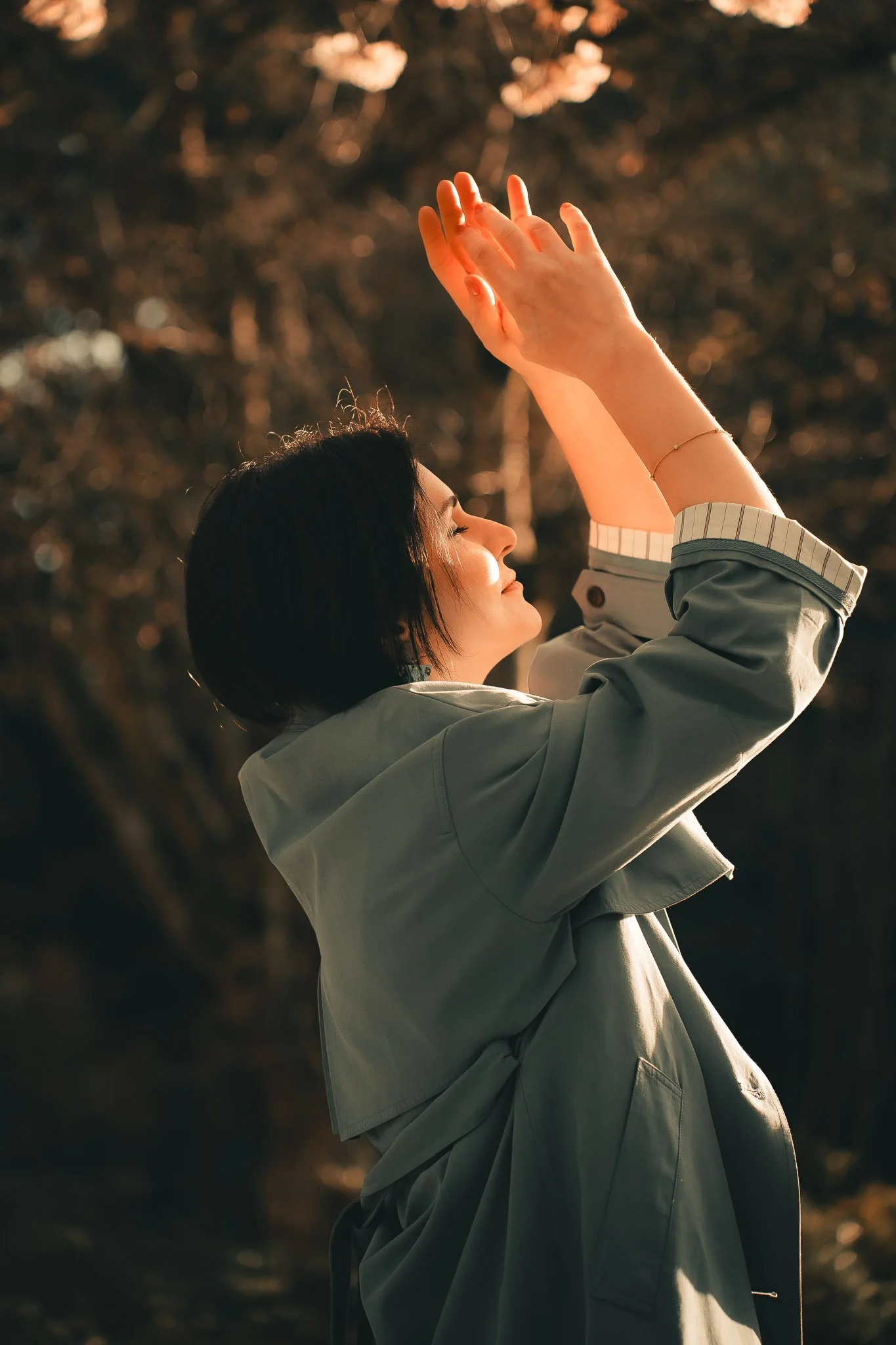 A woman with short dark hair in a green jacket stands outdoors with her eyes closed and hands raised, appearing to enjoy the moment in natural light.