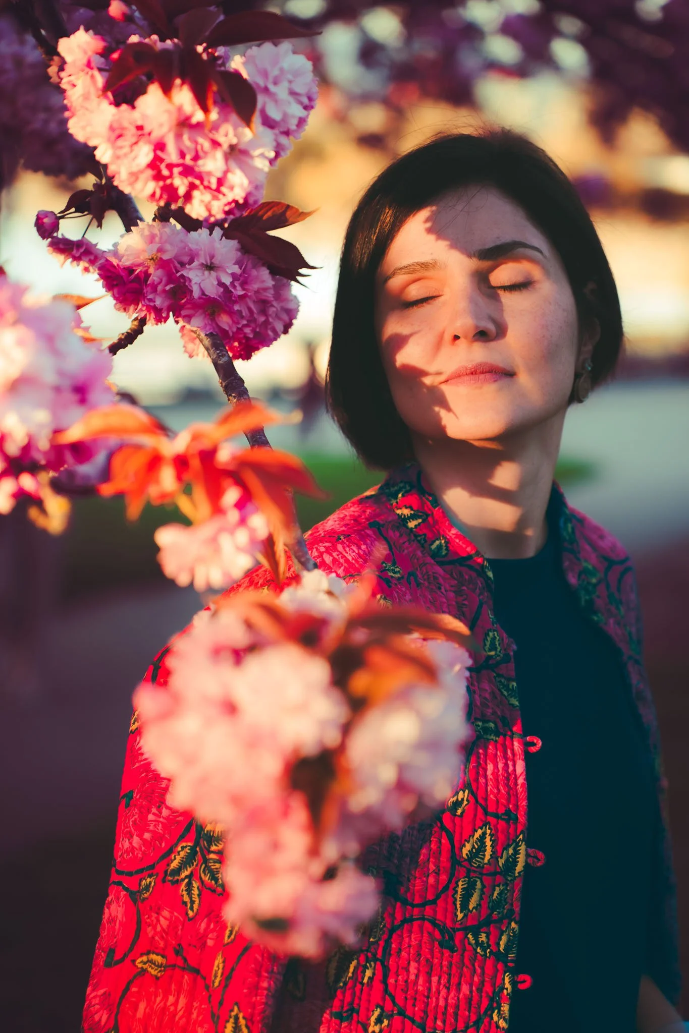 A woman with short black hair and closed eyes stands near pink and purple cherry blossoms during sunset, wearing a red and black patterned jacket.
