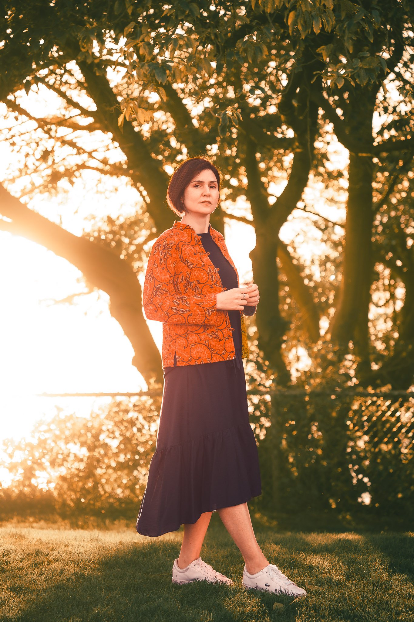 A woman stands outdoors during sunset in front of large trees, wearing a black dress, white sneakers, and an orange jacket with a leaf pattern.