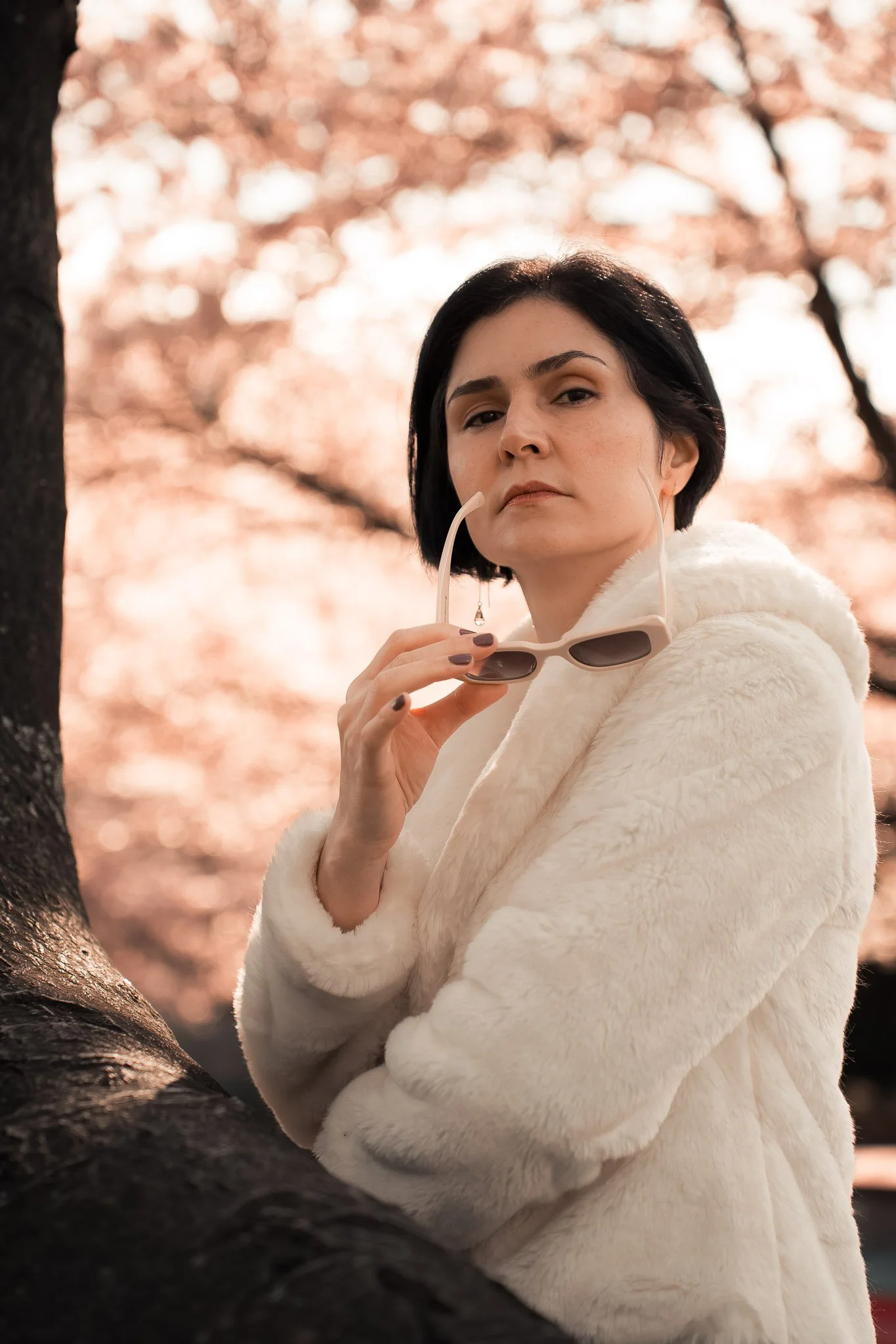 A woman with dark hair and light skin holding sunglasses stands near a tree with pink blossom background.