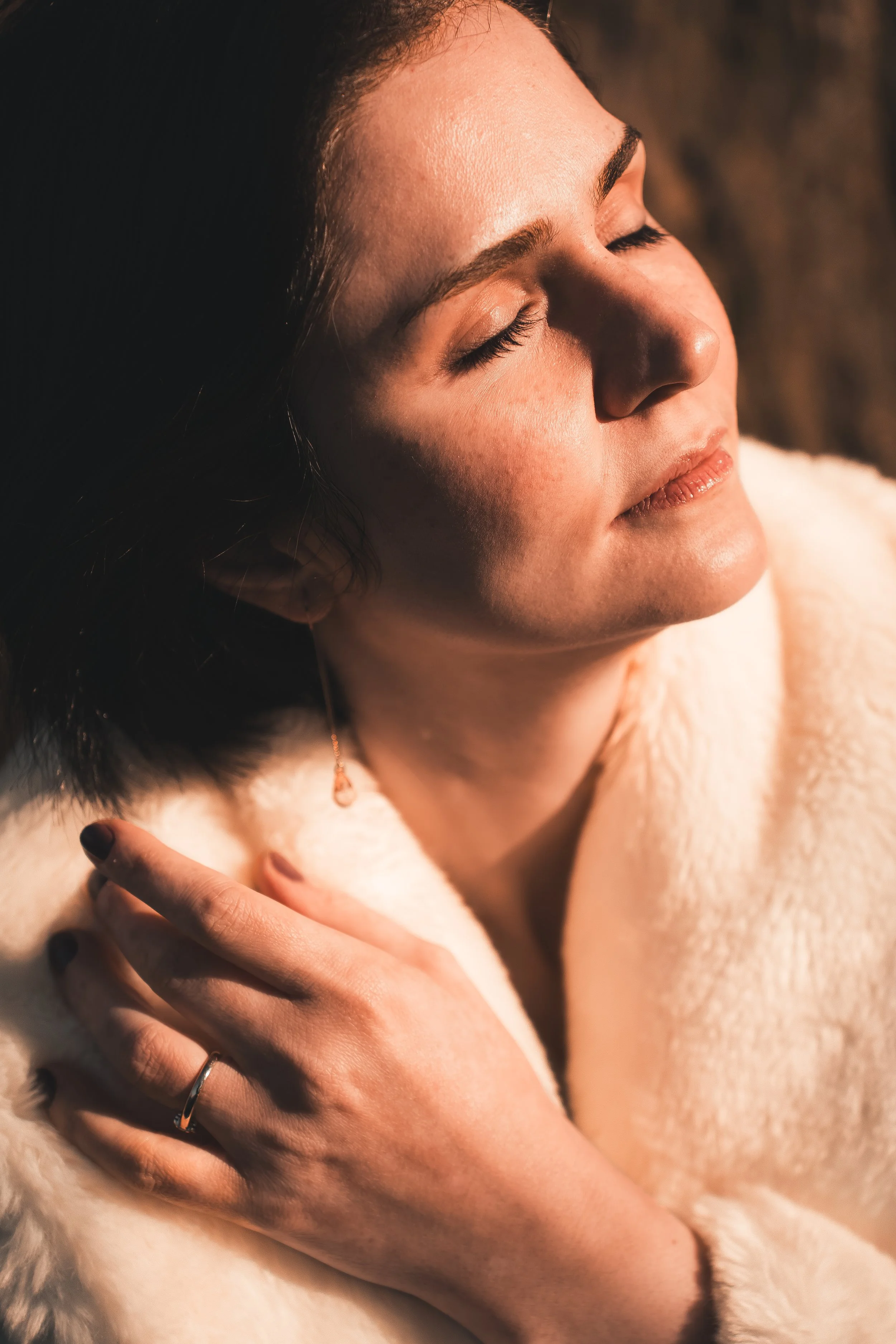 Close-up of a woman with dark hair and closed eyes, wearing a white furry garment, with her hand gently touching her shoulder.