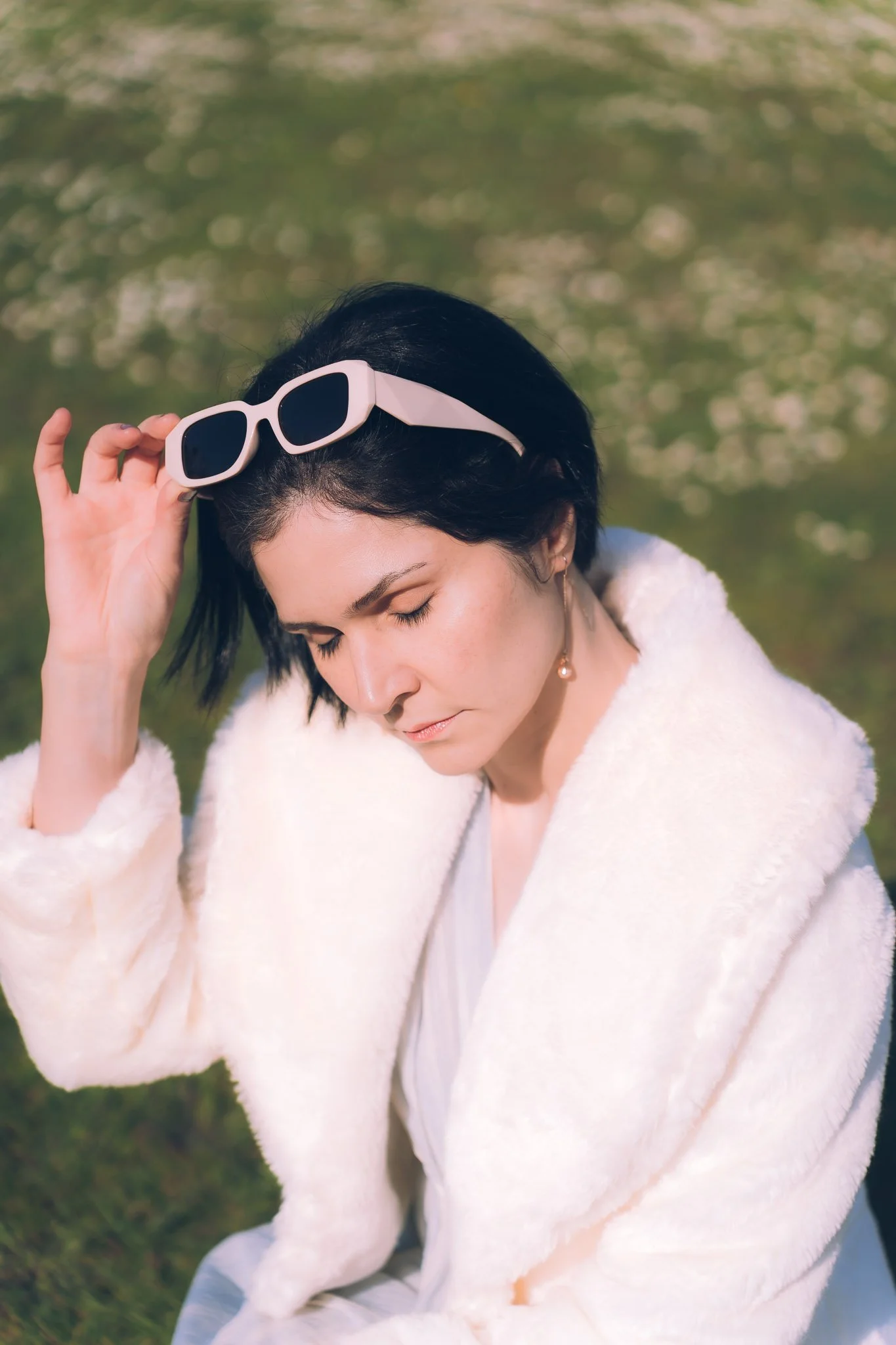 Woman with short black hair and light skin, wearing a white fluffy coat, sitting outdoors on grass, holding a pair of white sunglasses, with closed eyes.
