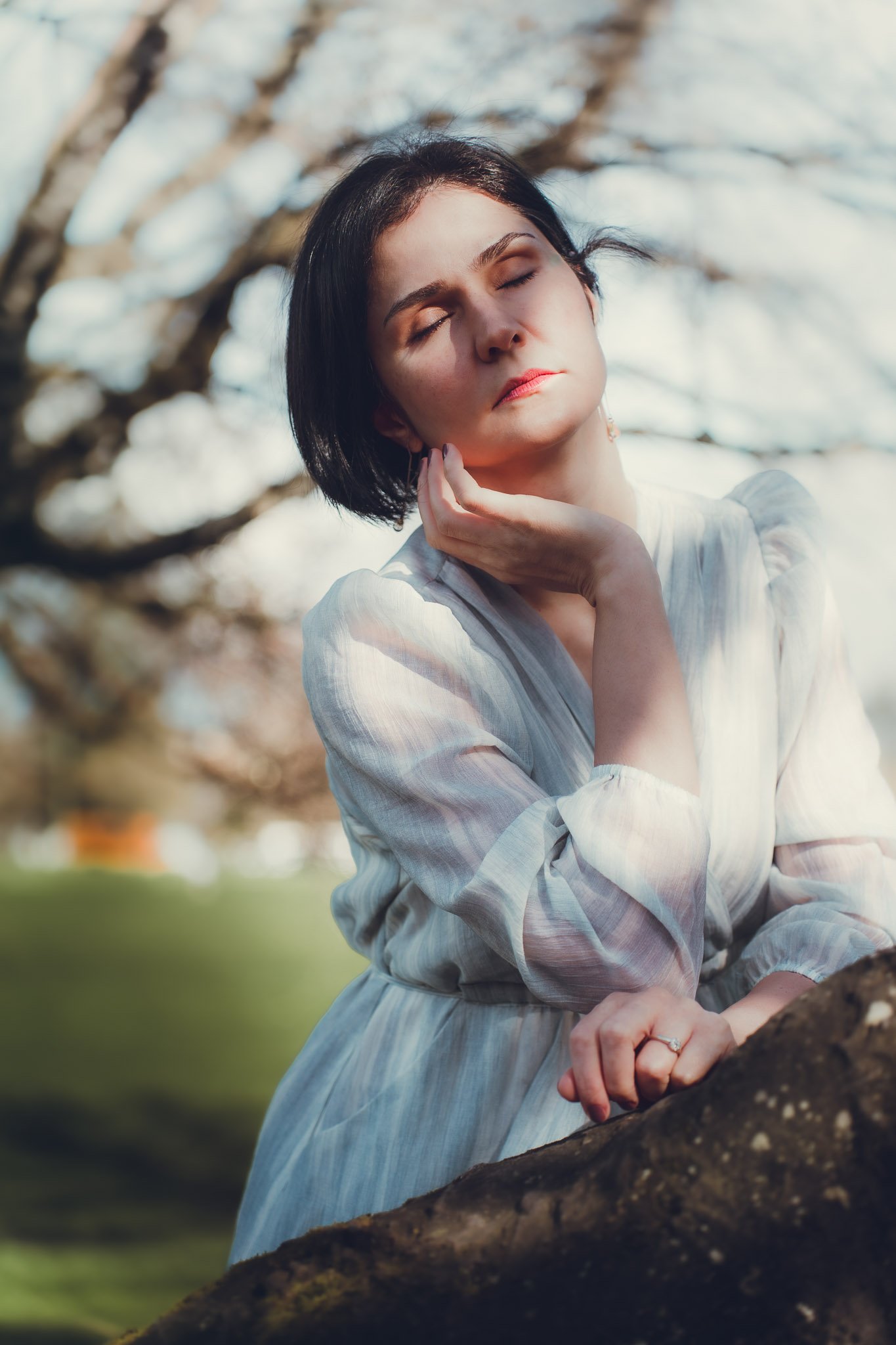 A woman with short dark hair and light skin, wearing a light-colored, striped dress, is outdoors with trees in the background. She has her eyes closed, her left hand gently touching her neck, and she appears to be in a peaceful, contemplative moment.