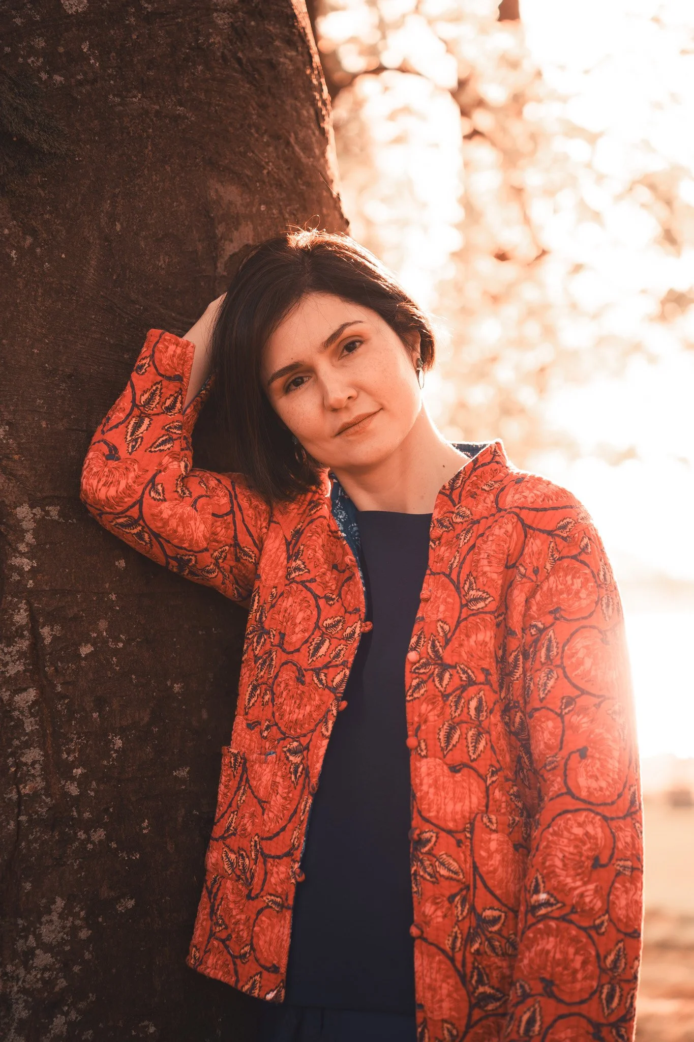 A woman with short dark hair leaning against a tree outdoors in warm lighting, wearing a patterned orange jacket over a dark shirt.