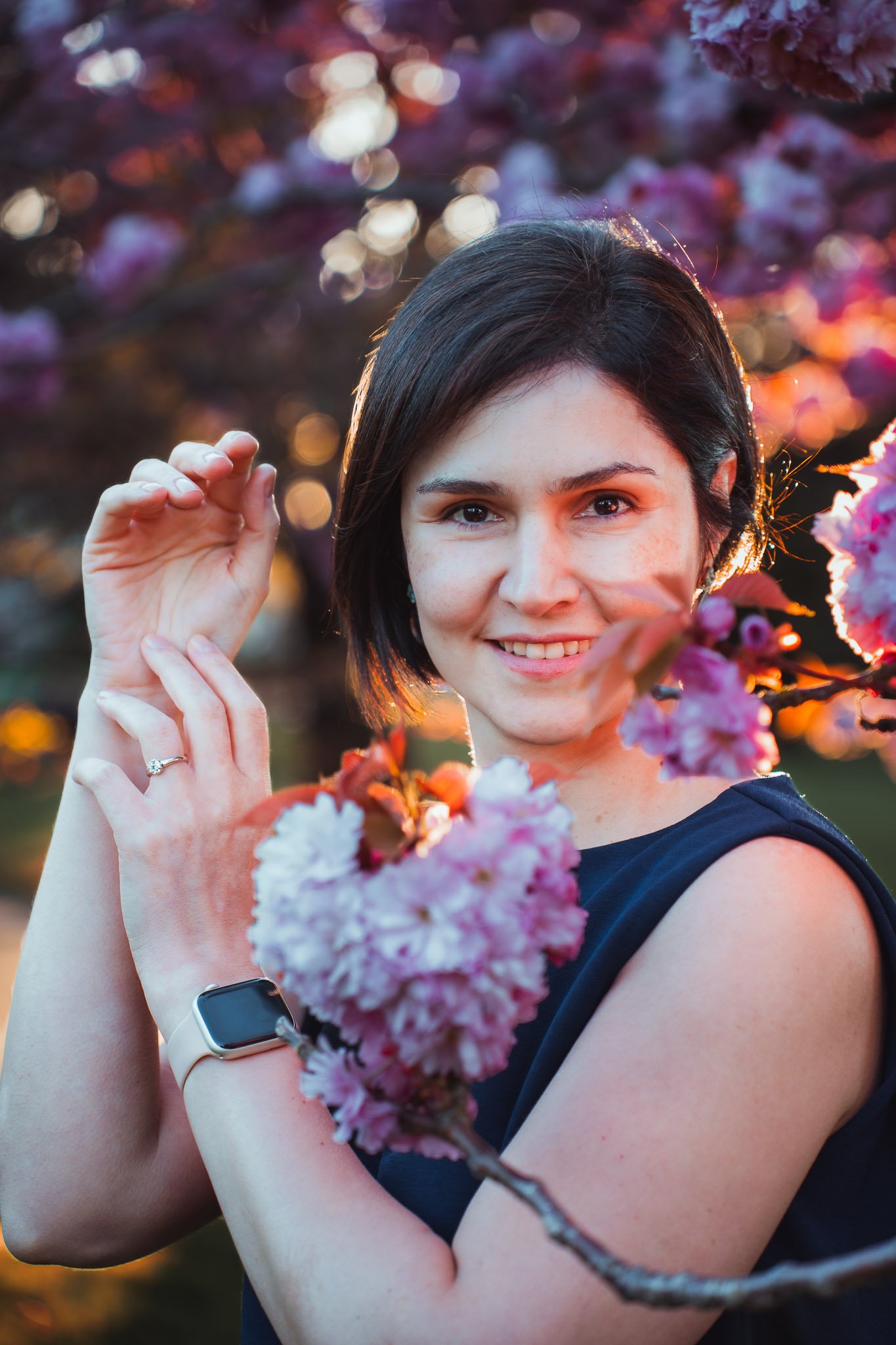 A woman with short dark hair smiling, standing near pink cherry blossoms during sunset, wearing a black sleeveless top, a smartwatch, and a ring on her finger.