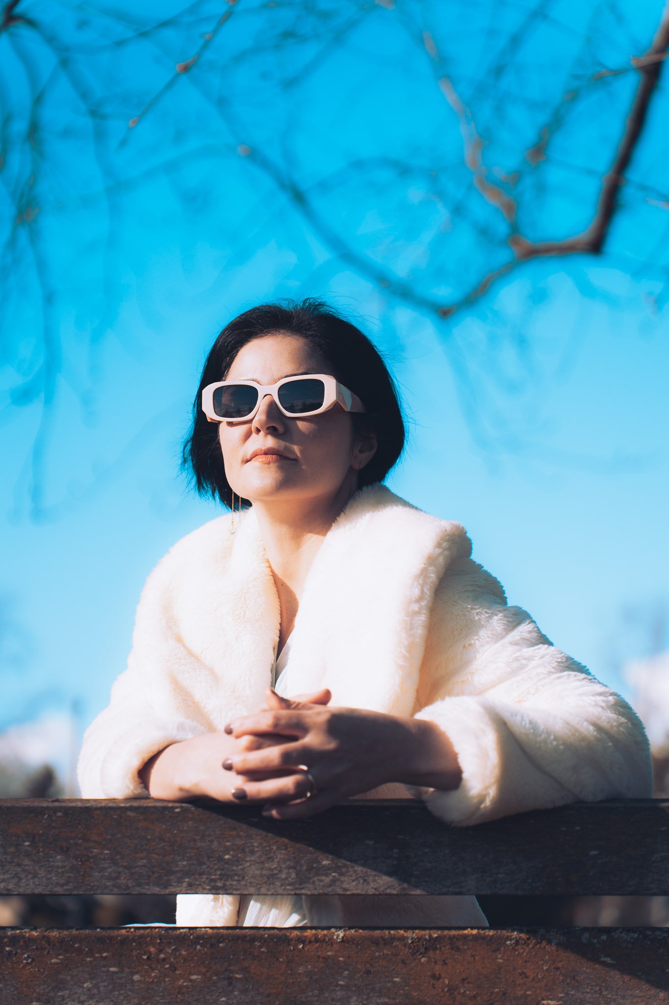 Woman wearing white fur coat and large sunglasses sitting at a wooden bench outside on a sunny day with blue sky and tree branches overhead.