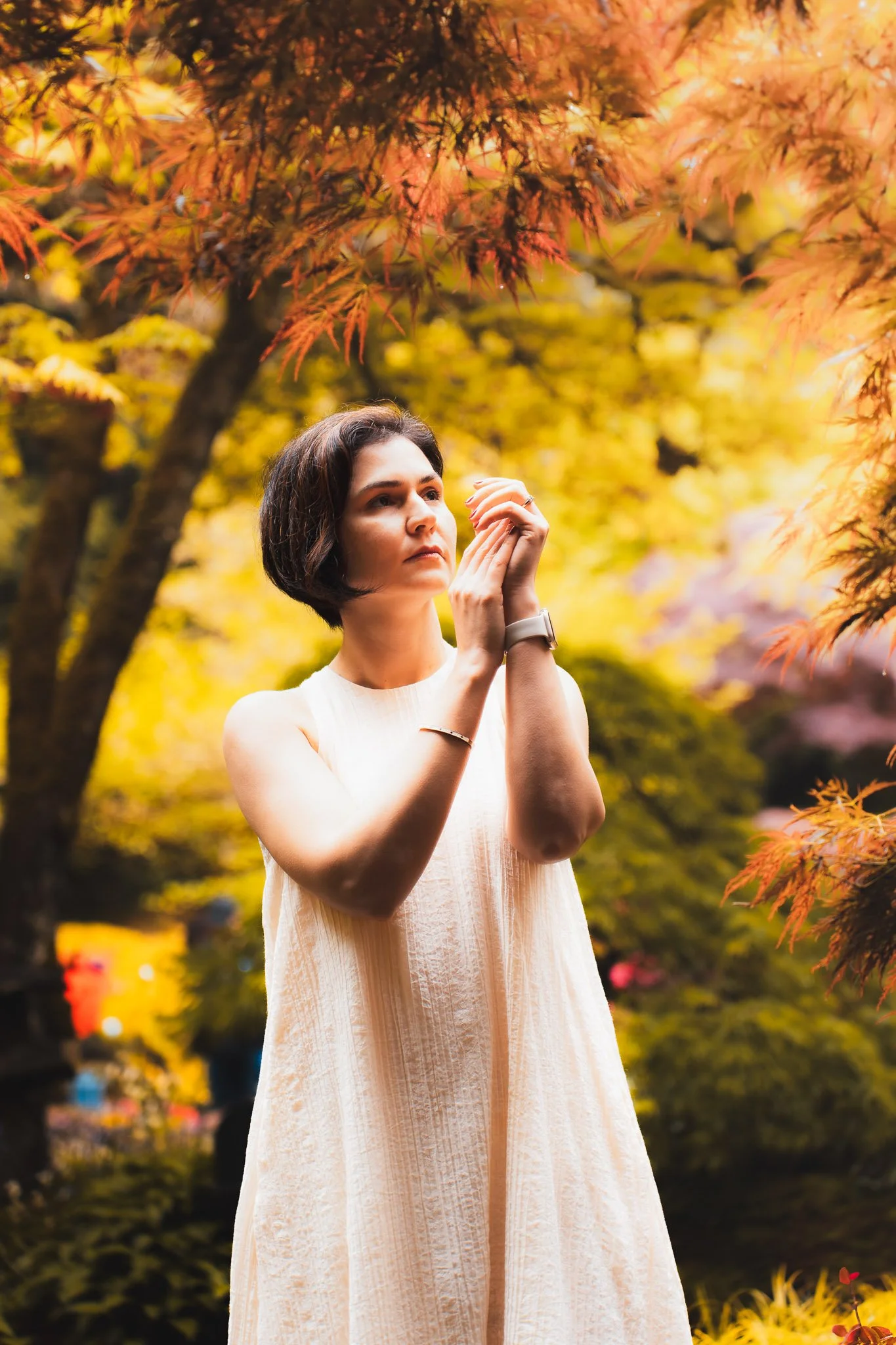 A woman with short dark hair standing outdoors among colorful autumn trees, wearing a sleeveless pale pink or beige dress, holding her hands clasped near her face, gazing thoughtfully into the distance.