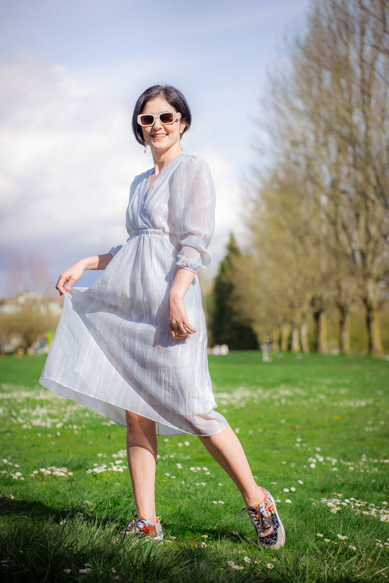 A woman with short dark hair, wearing sunglasses and a white dress, standing on a grassy field with trees in the background under a partly cloudy sky.