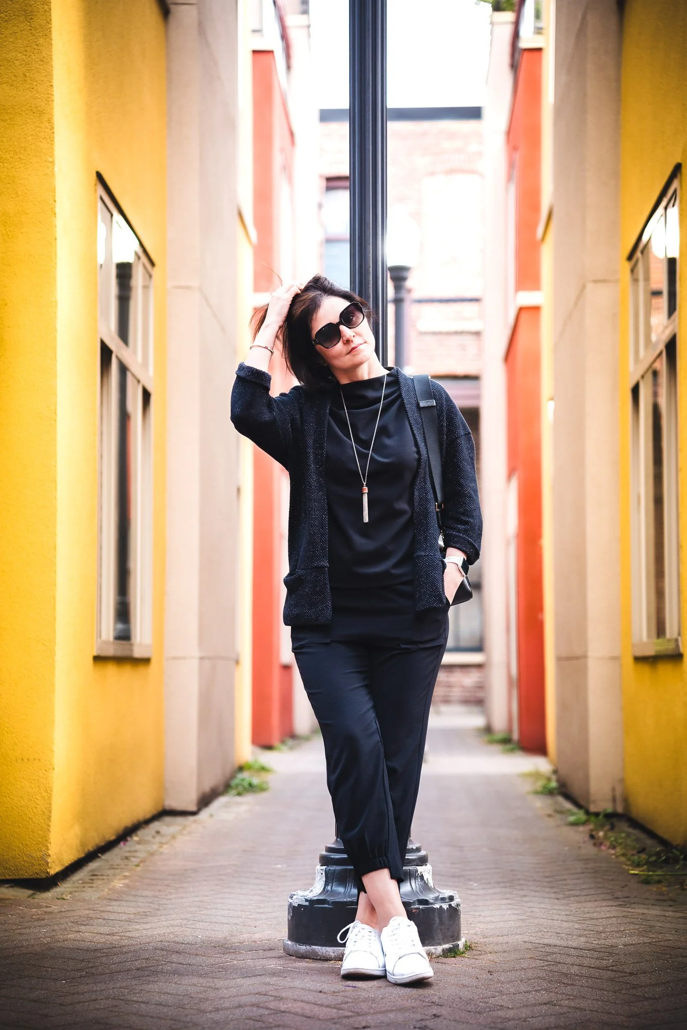 A woman dressed in black, wearing sunglasses, and white sneakers, standing in a colorful alley with yellow and orange walls, leaning against a street lamp.