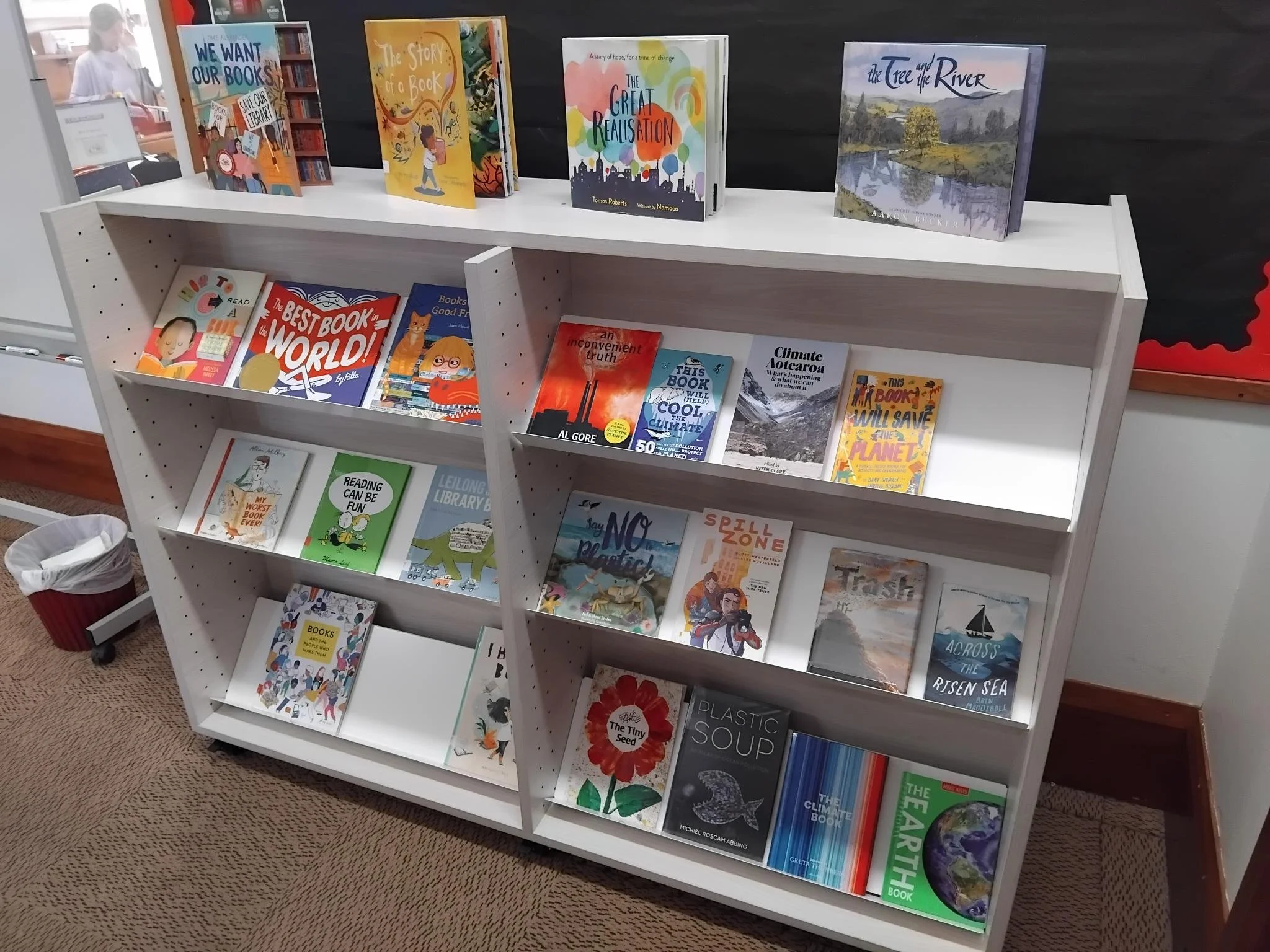 A white bookshelf with three shelves displaying various books. The top shelf features five children's books with colorful covers, including titles such as 'We Want Our Books!', 'The Story of a Book', 'The Great Realisation', 'The Tree and the River'. The middle shelf contains more children's books, including 'The Best Book in the World!' and 'Climate Ako,' among others. The bottom shelf displays additional books, some with nature-themed covers and titles related to the Earth and environment.