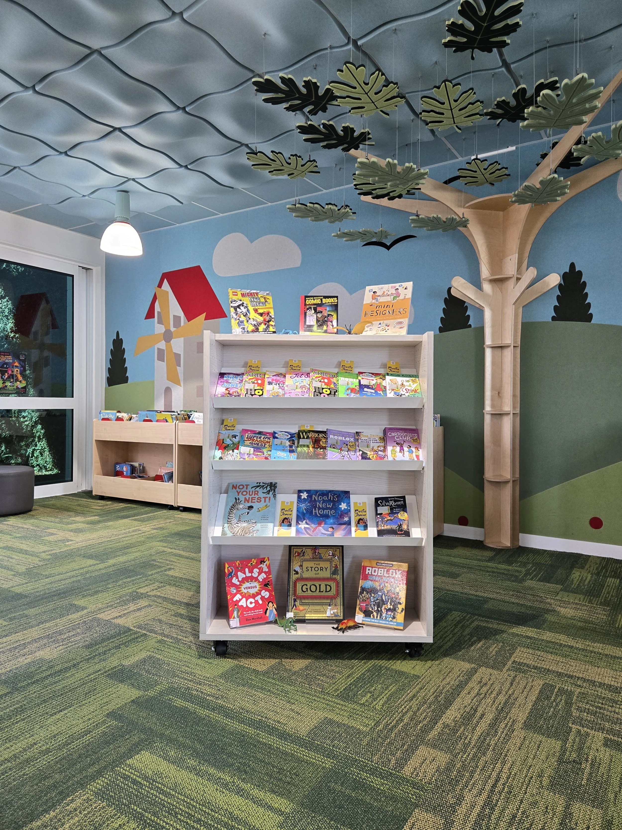 Indoor children's bookstore with a white book rack displaying children's books, decorated with a painted mural of a windmill, trees, clouds, and birds, and a large wooden tree structure with hanging leaf cutouts on the right side.