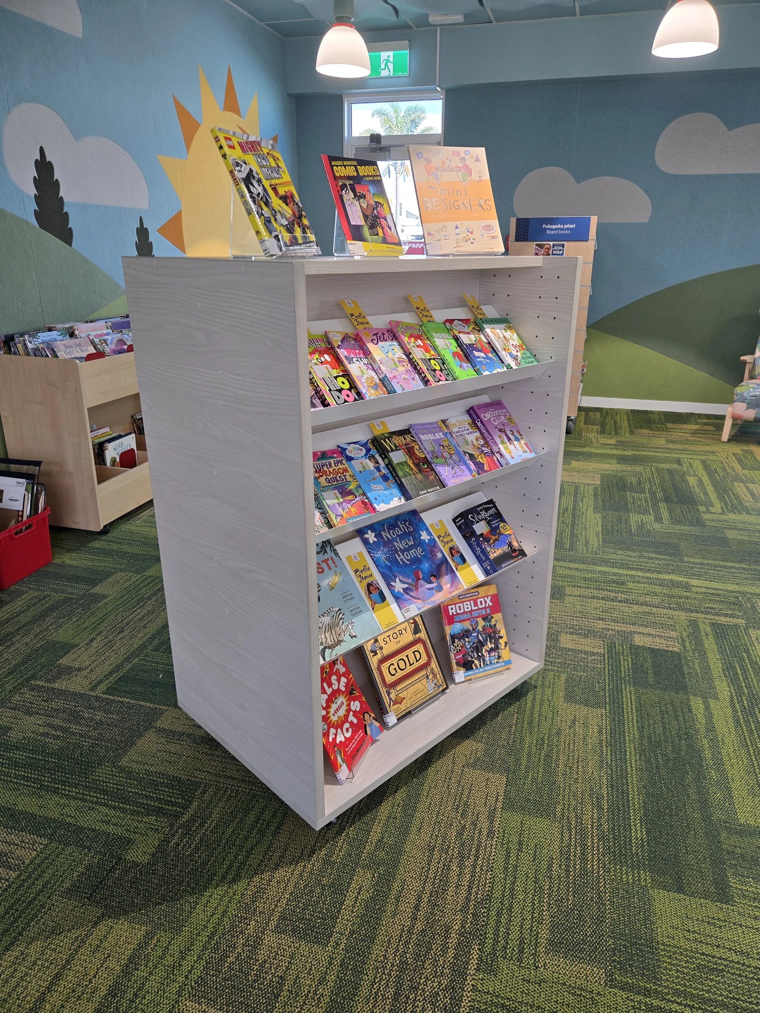 A white book display stand with colorful books on it inside a children's library or bookstore. The background has a painted mural of a sun, clouds, and green hills.