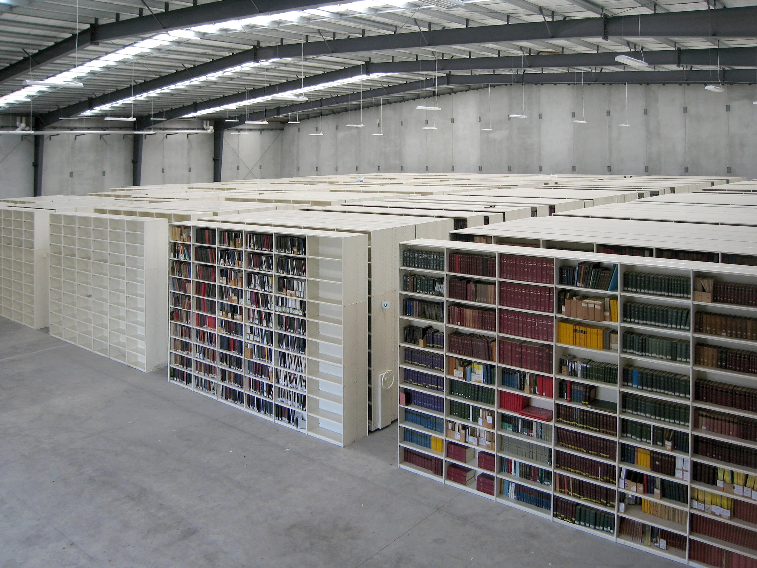 Interior of a large industrial warehouse with high ceilings, gray walls, bright overhead lighting, and bookshelves filled with books arranged in rows.