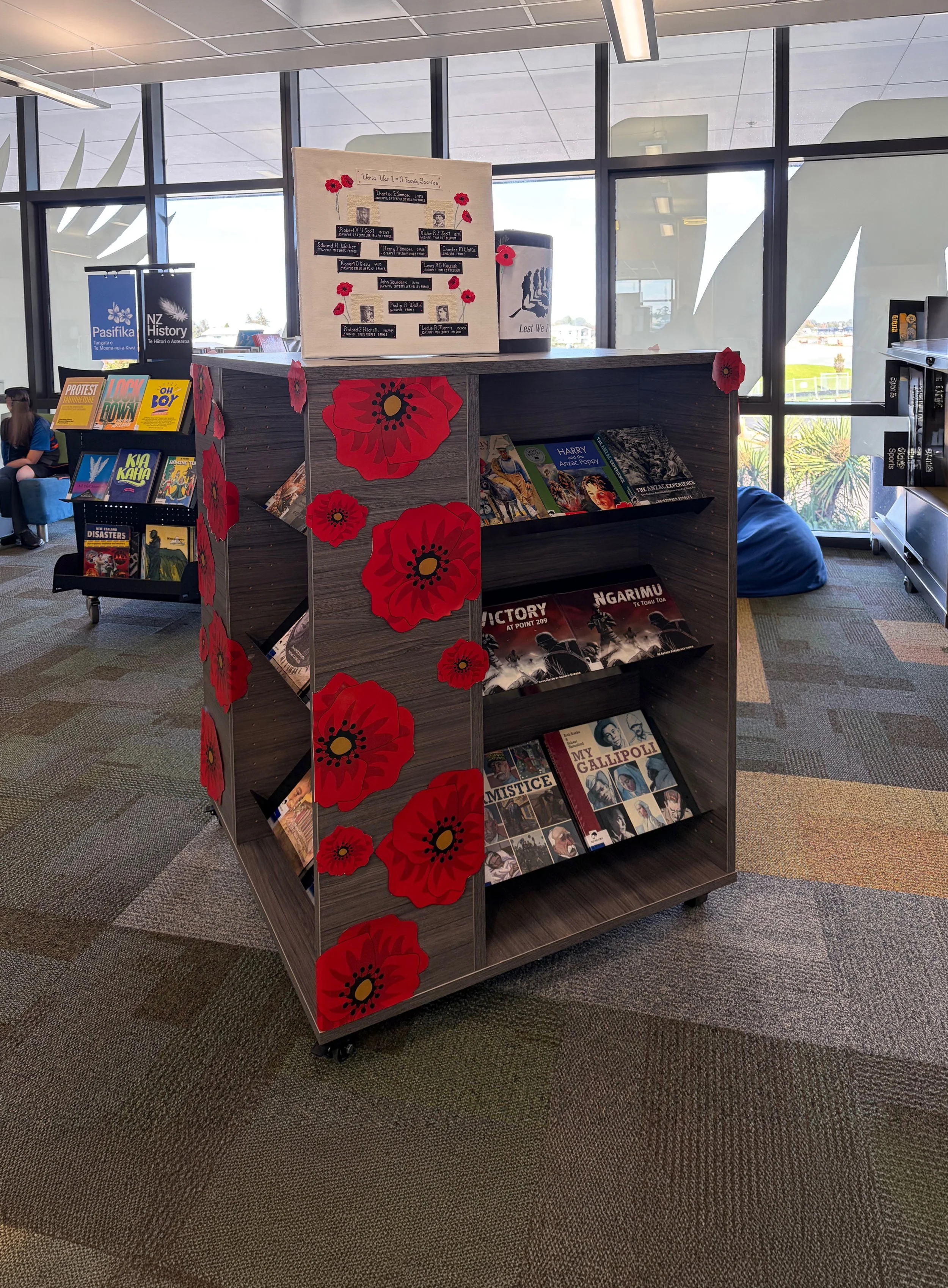 A wooden bookshelf decorated with red poppy flowers, containing various books on history and military topics, is located in a room with large windows and an outdoor view.