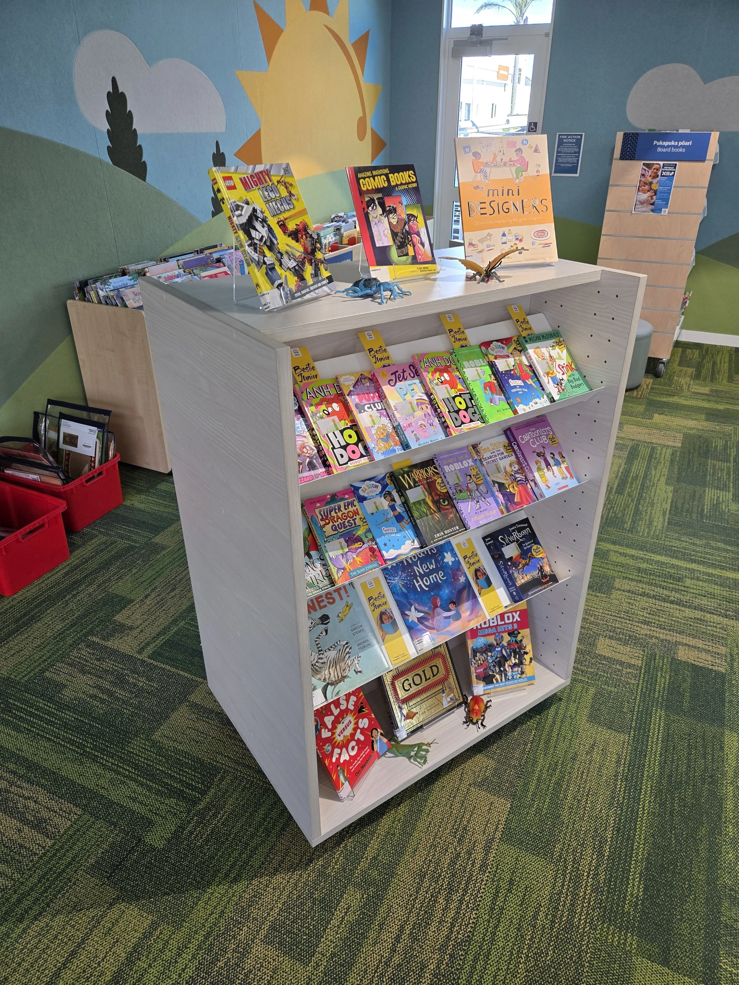 Book display shelf in a children's section of a library, featuring colorful books including comic books and storybooks, with a mural of a cartoon sun, clouds, and trees on the wall behind.