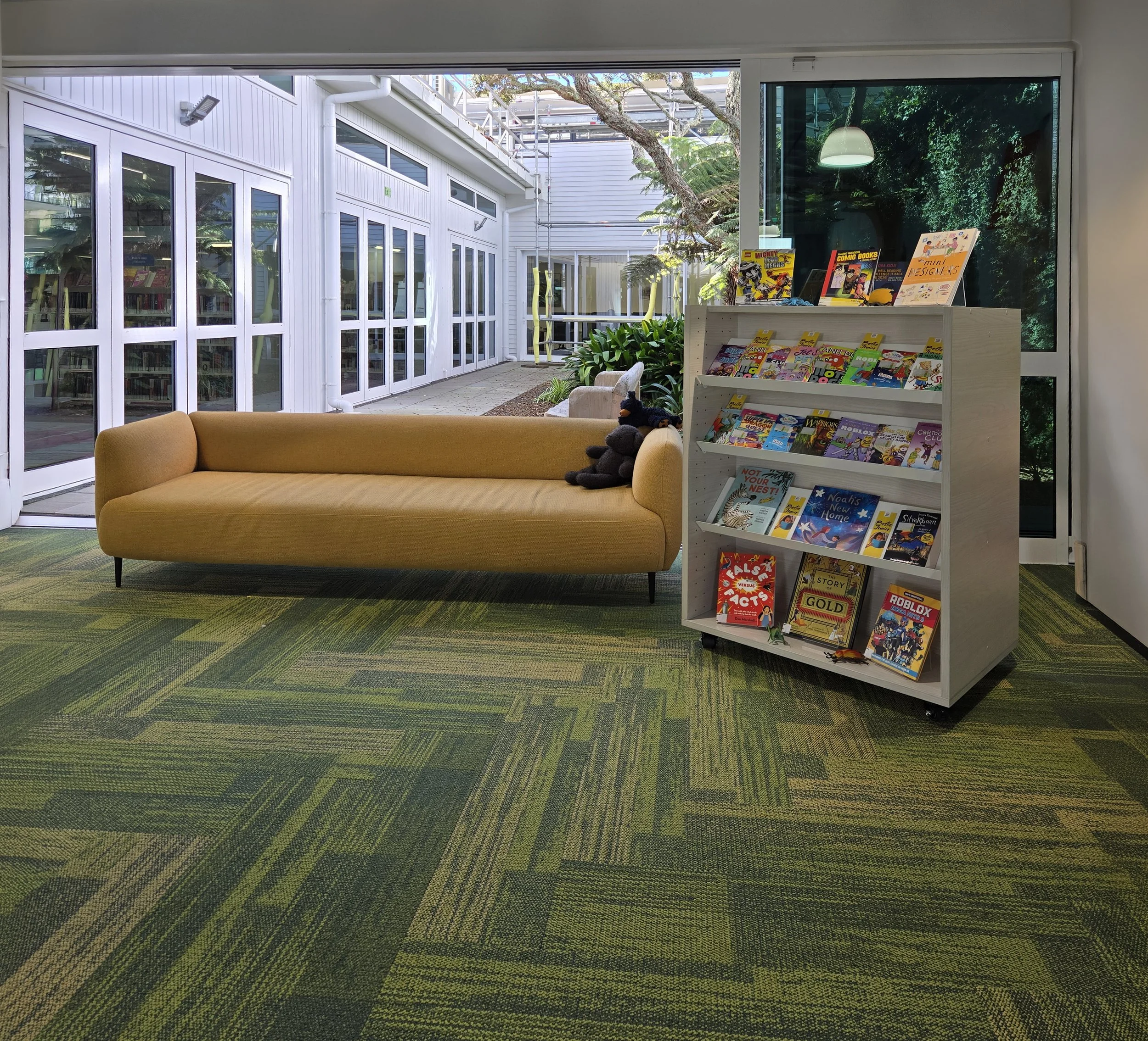 Indoor space with a yellow sofa, a white bookshelf filled with comic books, and full-length glass windows revealing an outdoor corridor with plants and a building.