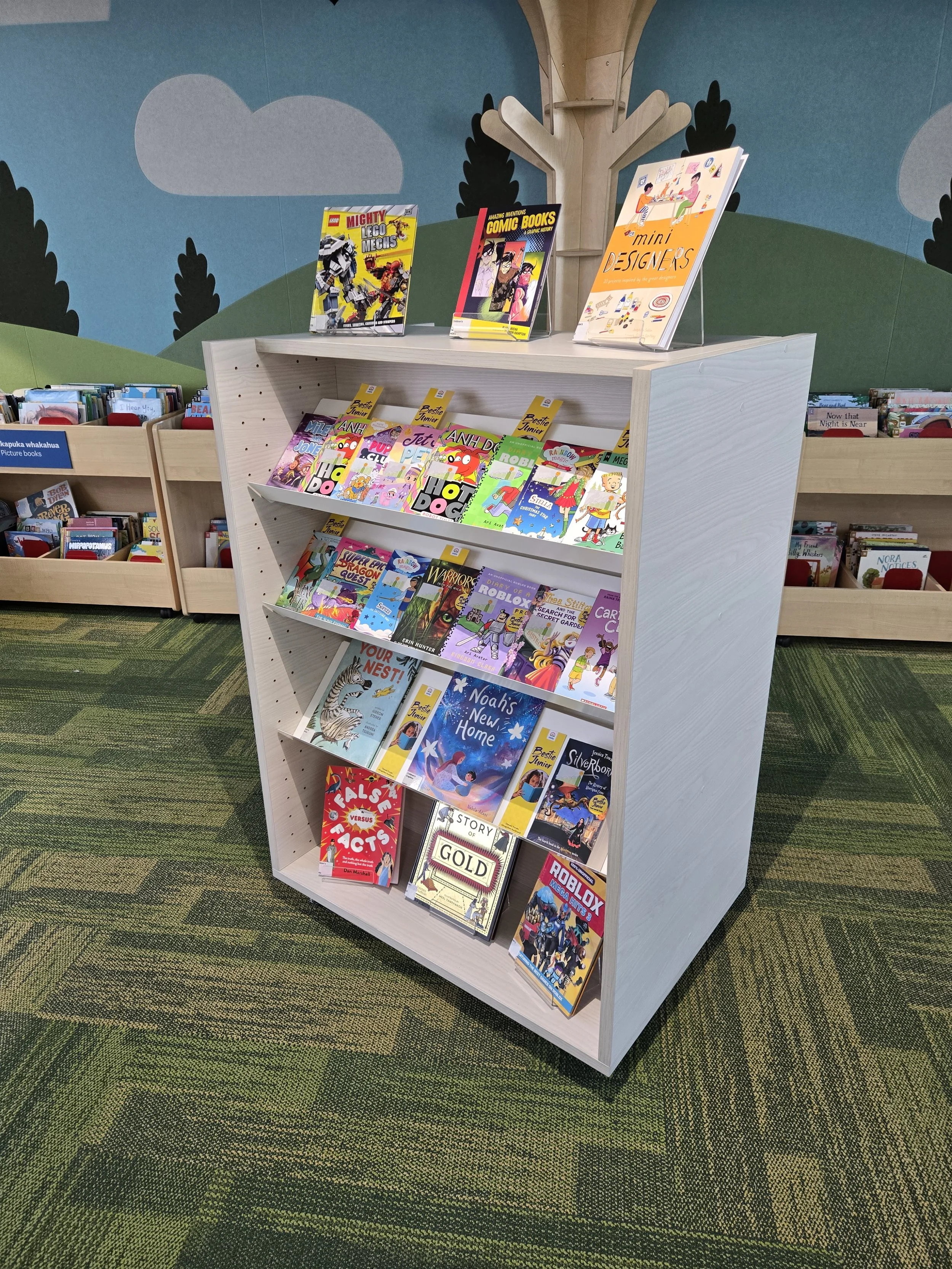 Book display in a library featuring children's books, with colorful covers, on a white bookshelf. The background has a painted mural with clouds, trees, and hills.